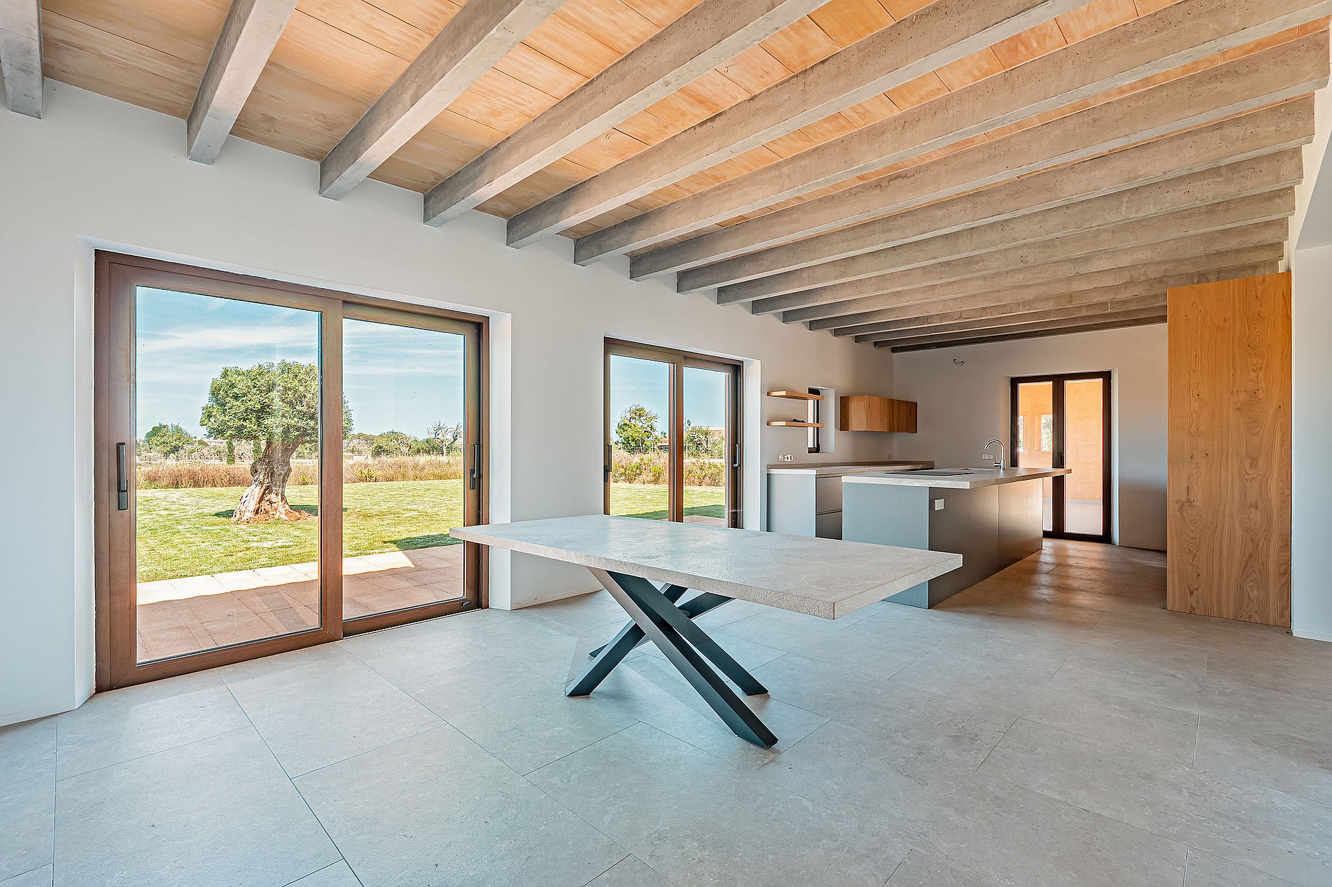 Dining area with a stone table and dark metal legs, exposed wooden ceiling beams, and large glass doors overlooking a rural landscape.