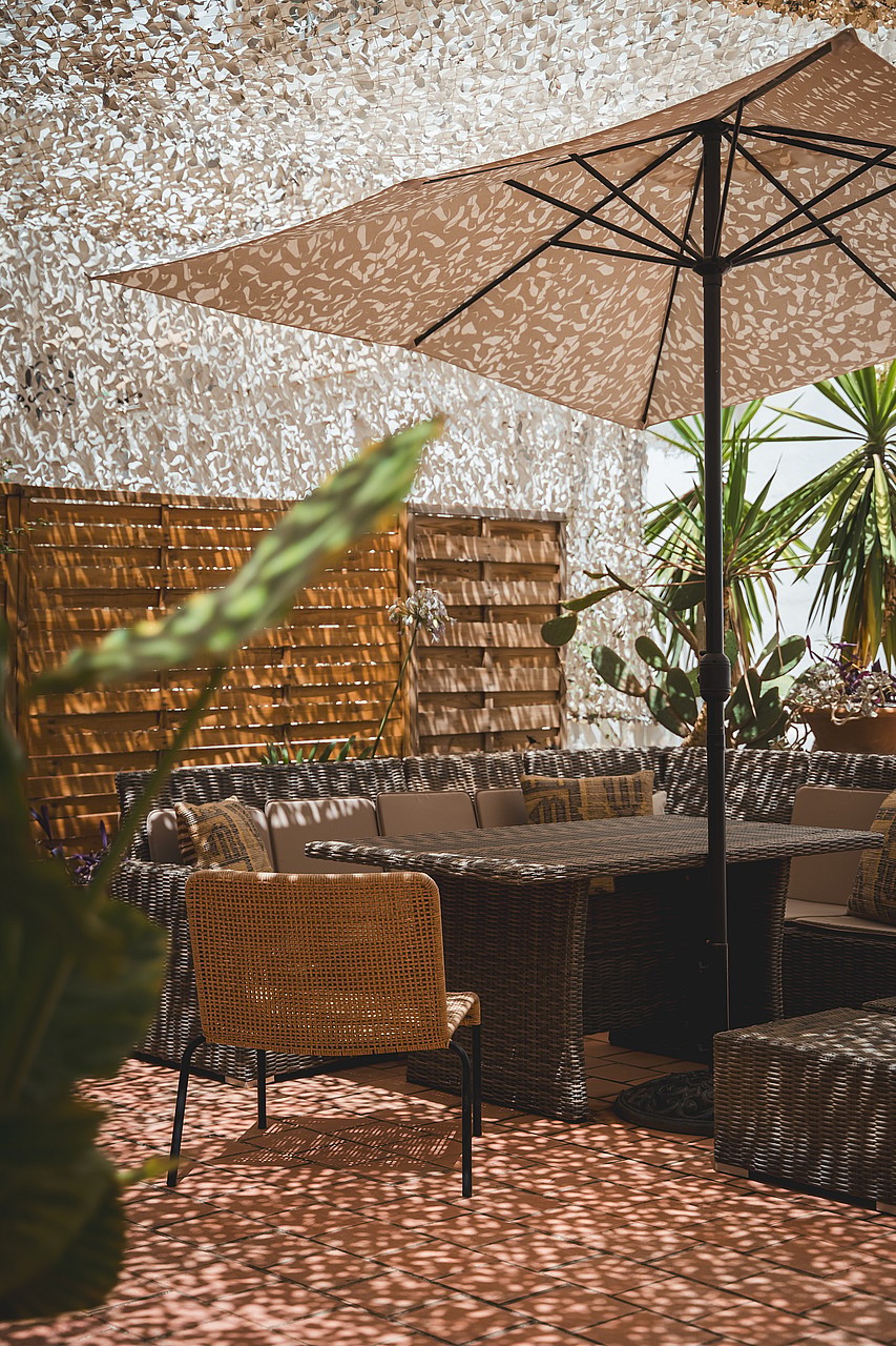 A shaded outdoor terrace with a dining table and rattan chairs, featuring a camouflage net canopy and wooden pallet screens.