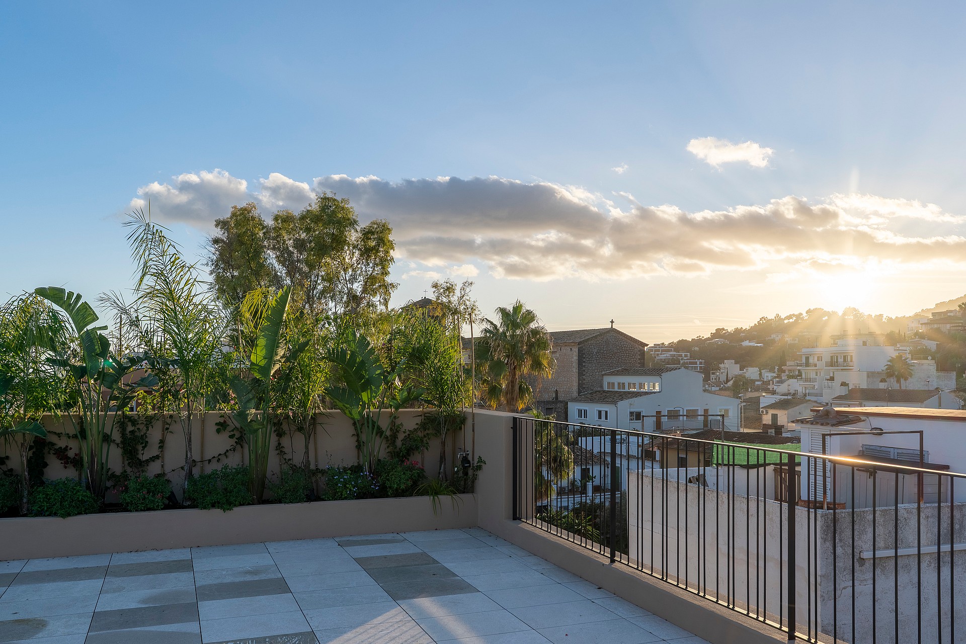 A stone-paved rooftop terrace featuring black metal railings and lush planters, overlooking village rooftops and hills at sunset.