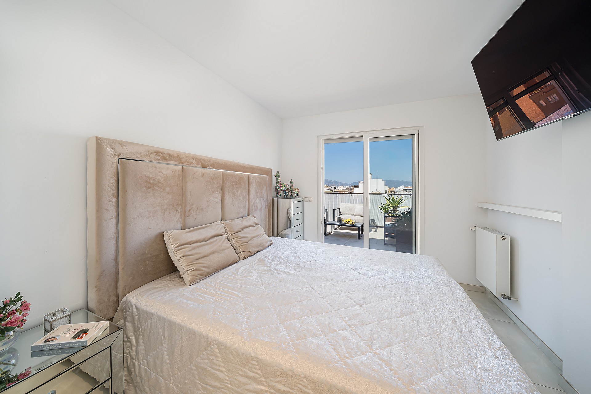 A bright bedroom with a queen bed, upholstered beige headboard, and a glass sliding door leading to an outdoor balcony.
