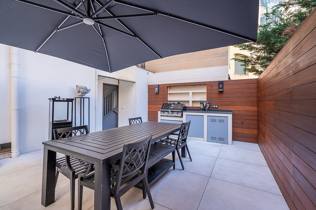 A private outdoor terrace in Palma featuring a charcoal grey dining set under a large umbrella, adjacent to a wooden-paneled summer kitchen.