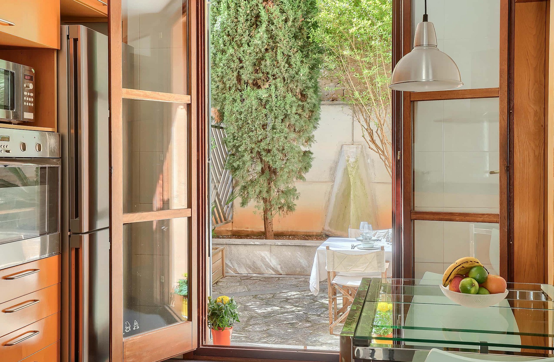 A kitchen interior featuring warm wooden cabinetry, a glass-topped table with fruit, and full-height glass doors leading out.