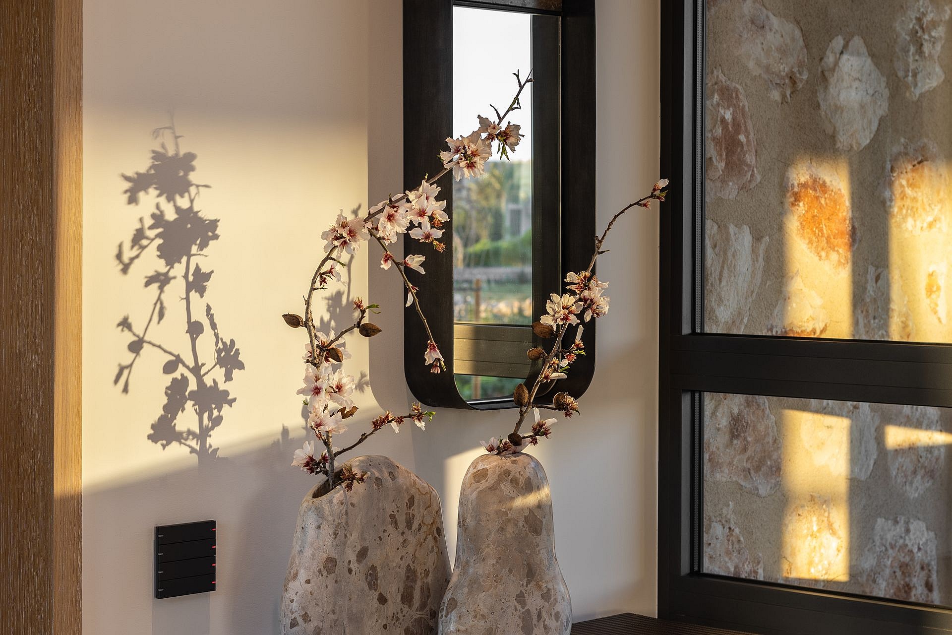 Close-up of flowering branches in stone vases on a surface near a stone wall and modern black-framed window.
