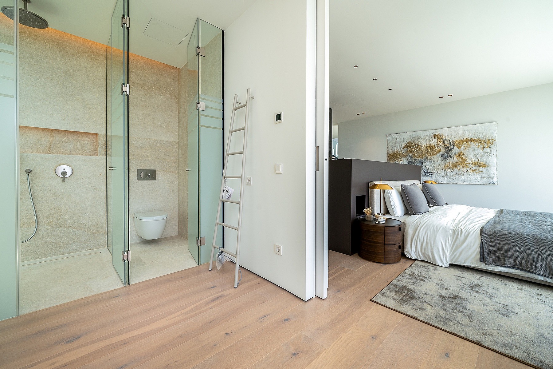 An open-plan bedroom with light wood floors and a glass-enclosed shower room, featuring modern fixtures and neutral tones.