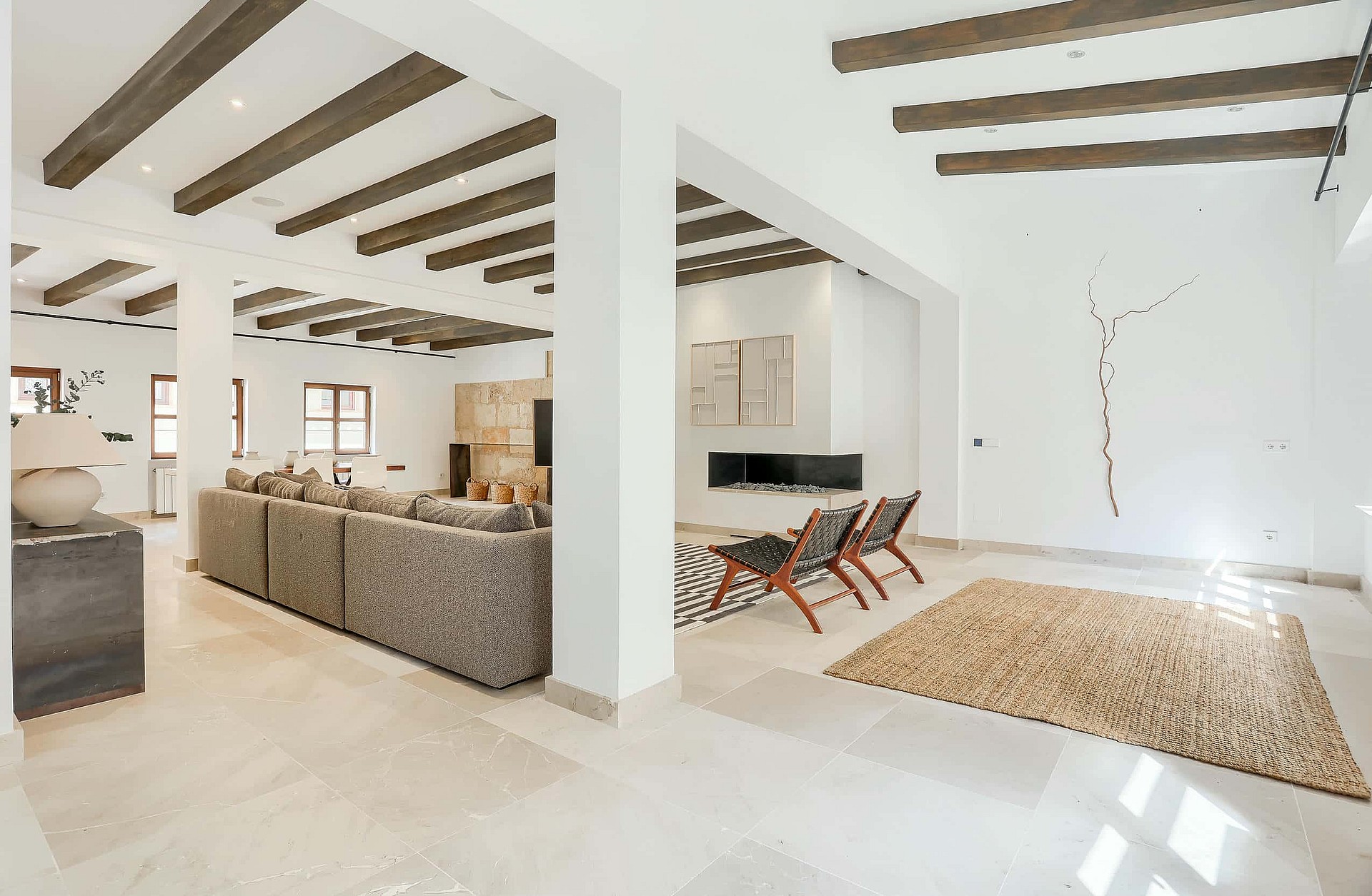 Interior view of a bright, spacious living room with white walls, exposed timber ceiling beams, stone flooring, and a large sofa.