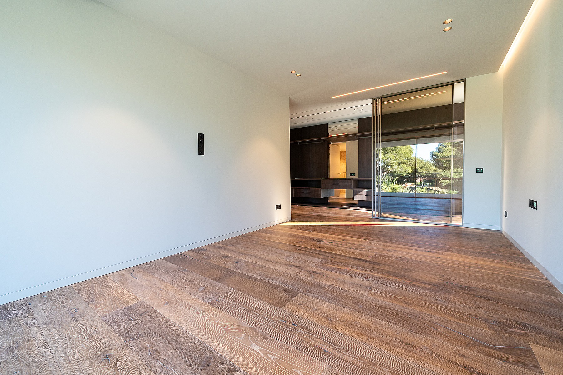 Empty interior room with wide-plank oak flooring and recessed ceiling lighting leading to a terrace with garden views.