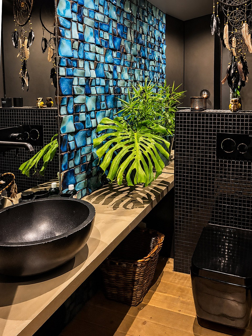 Interior of a bathroom featuring a bright blue mosaic tile wall, a black vessel sink, and a large monstera plant.