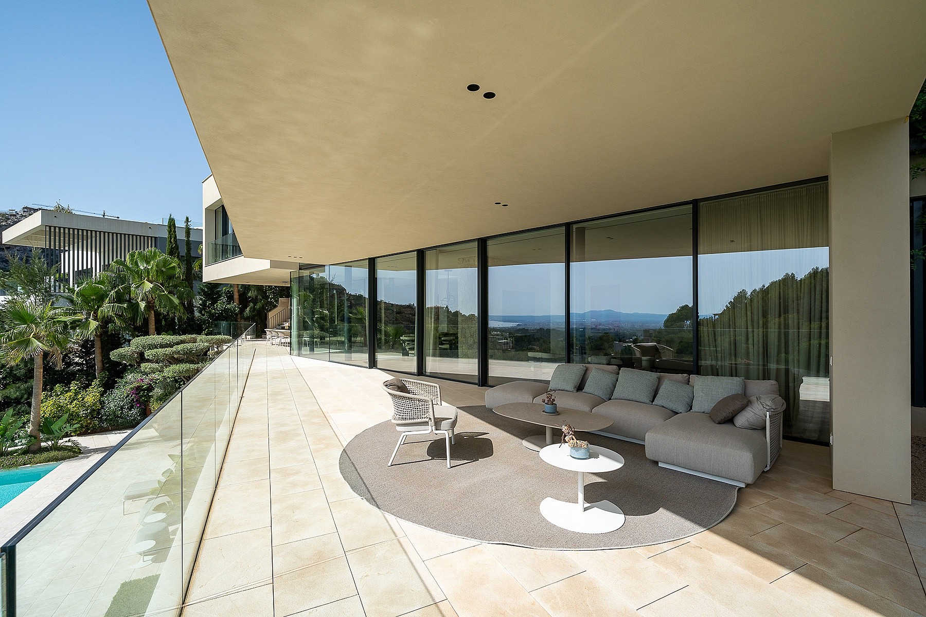 A wide, covered outdoor terrace with beige stone flooring, a modular sofa on a circular rug, and glass balustrades.
