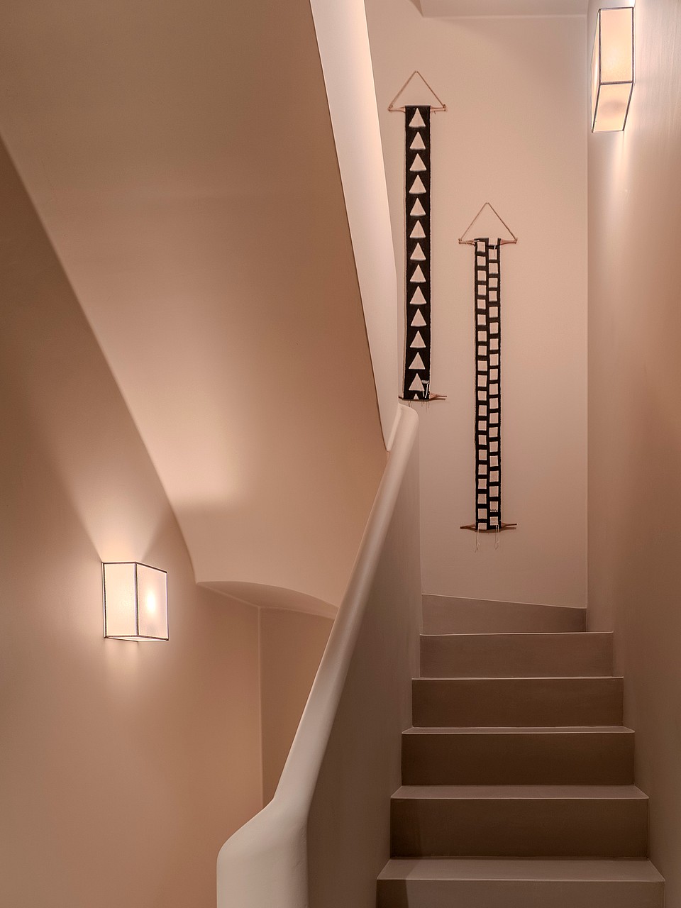 Modern staircase with smooth plaster walls, warm wall sconces, and vertical geometric wall art.