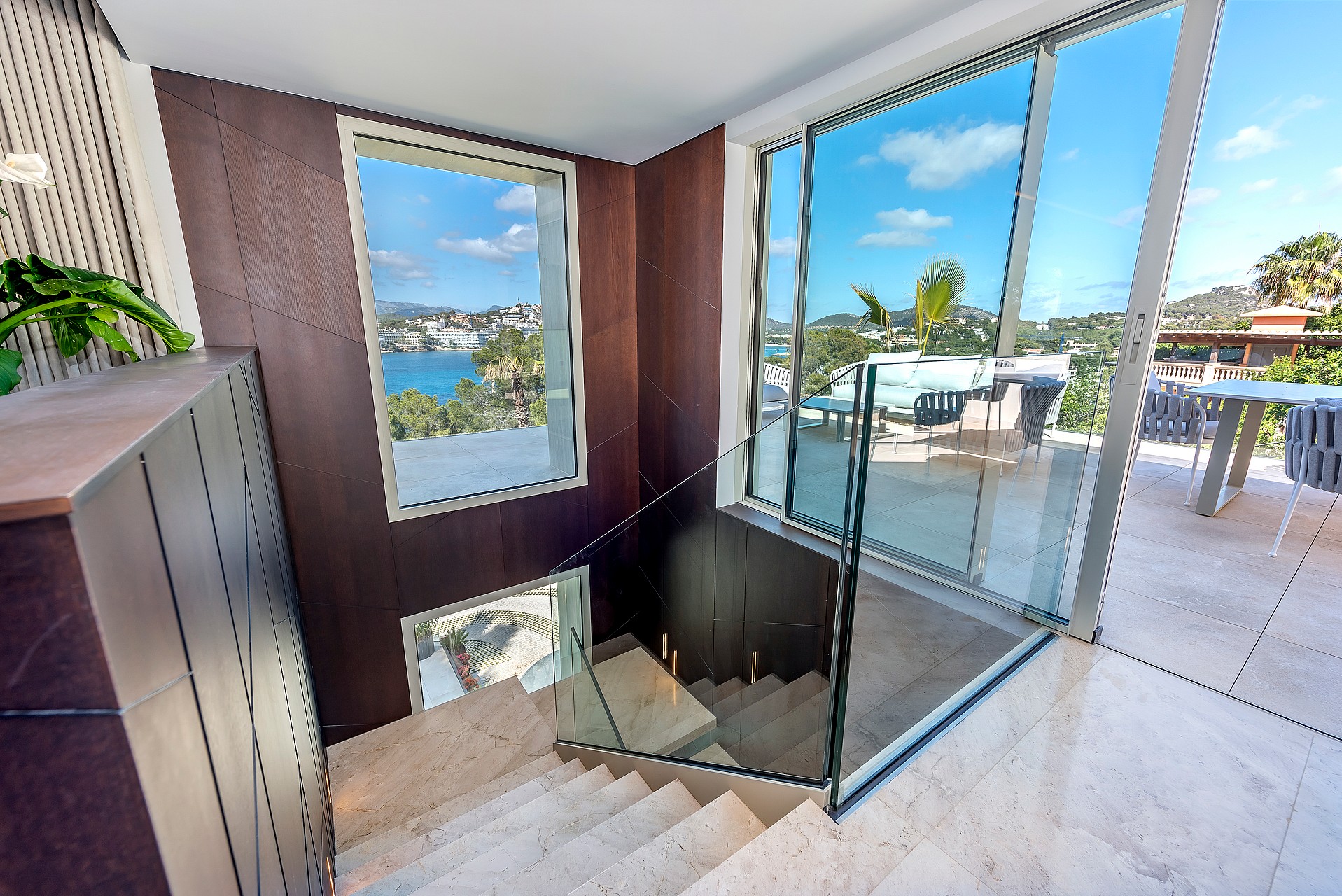A high-angle view looking down a marble staircase with a dark wooden feature wall, glass balustrades, and large windows looking out toward a blue bay.