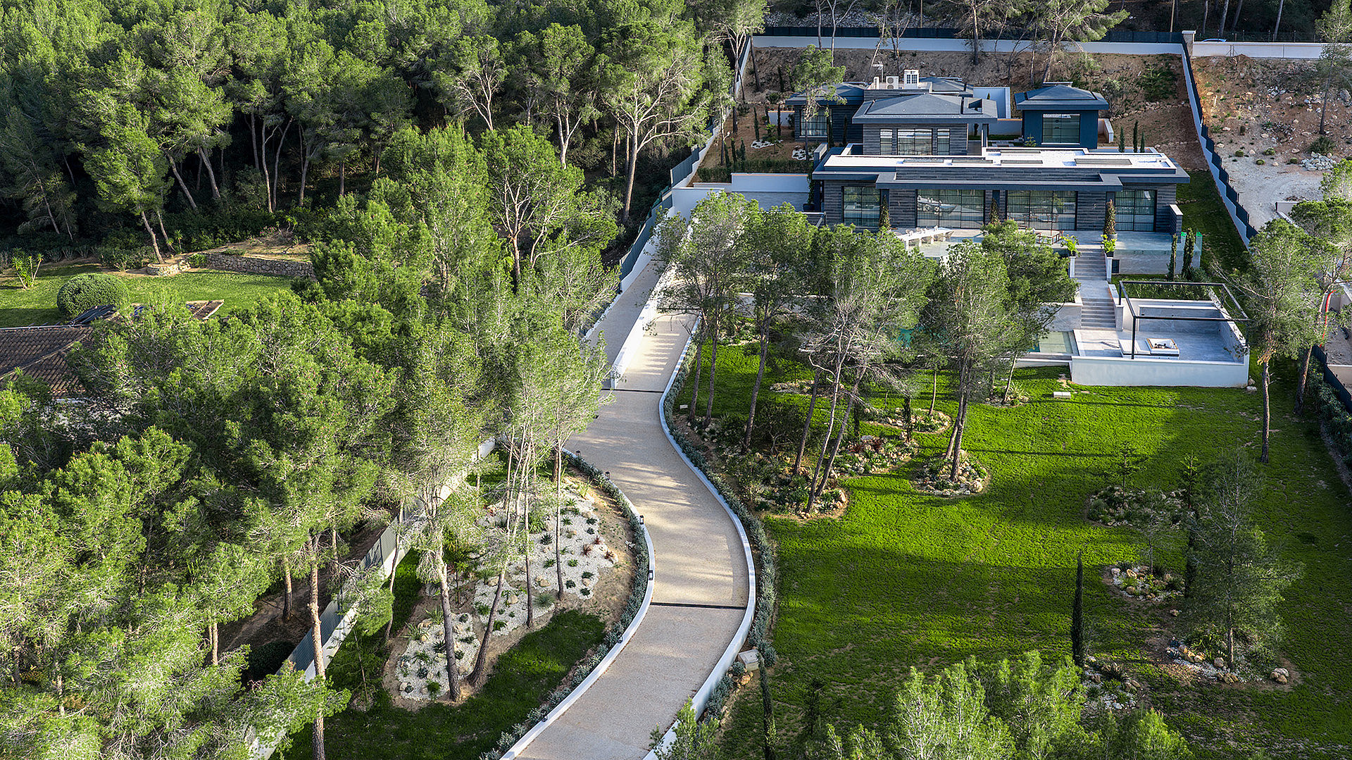 Aerial view of a modern villa with a flat roof, surrounded by gardens, pine trees, and a winding driveway.