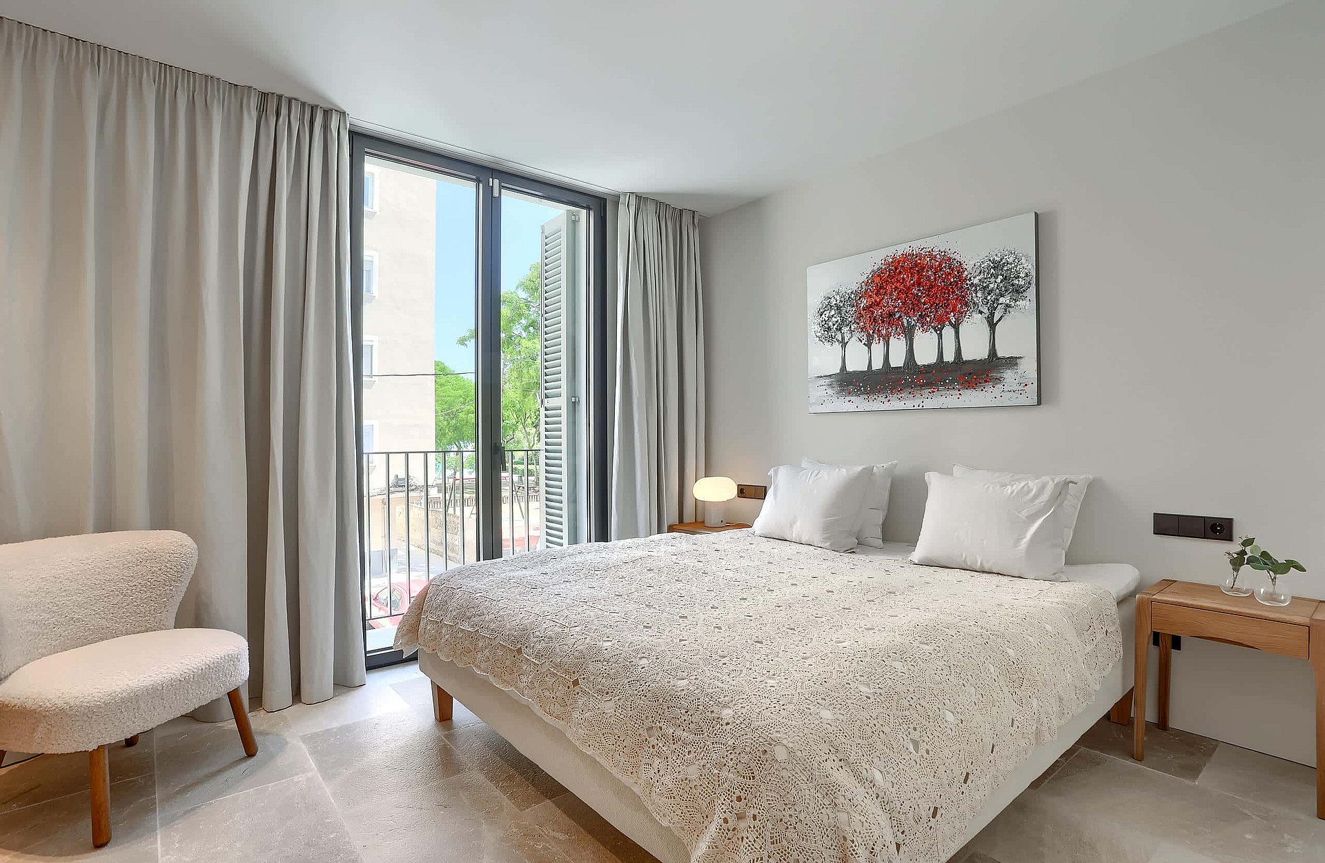 A contemporary bedroom featuring a queen bed with white bedding, a balcony door with grey curtains, and a textured armchair.