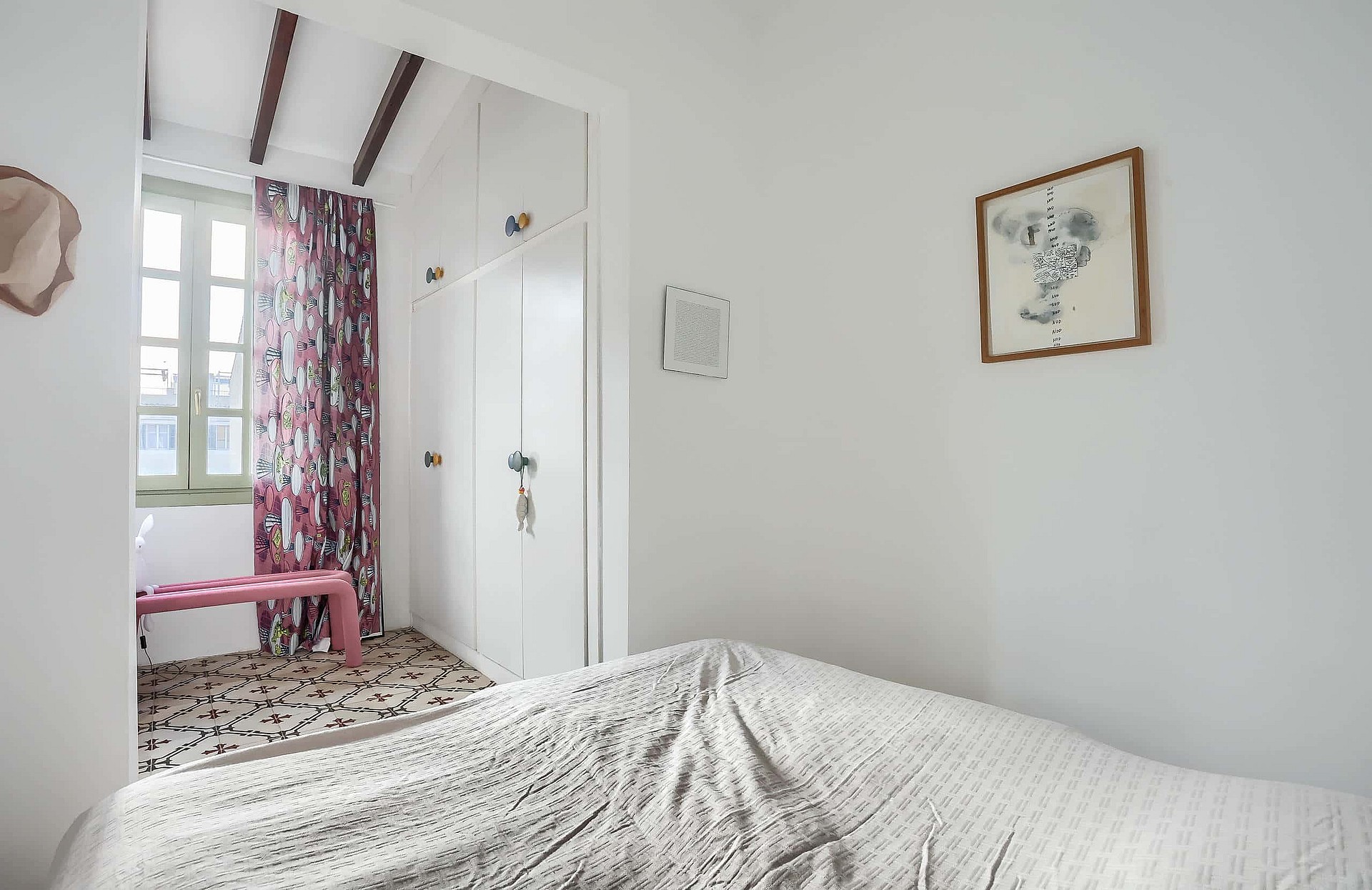 A bedroom featuring white built-in wardrobes, a patterned rug, and a pink bench near a window with floral curtains.