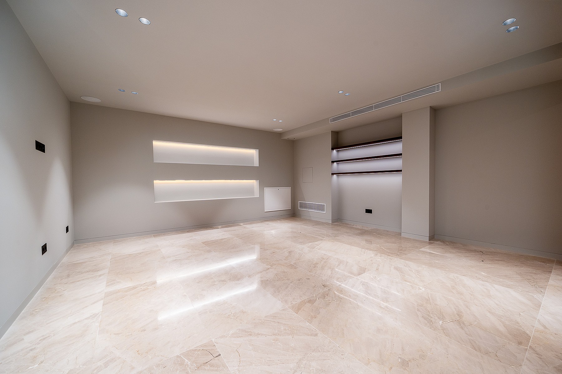 A spacious, unfurnished room with beige stone floors, recessed wall shelving with integrated LED lighting, and neutral walls.
