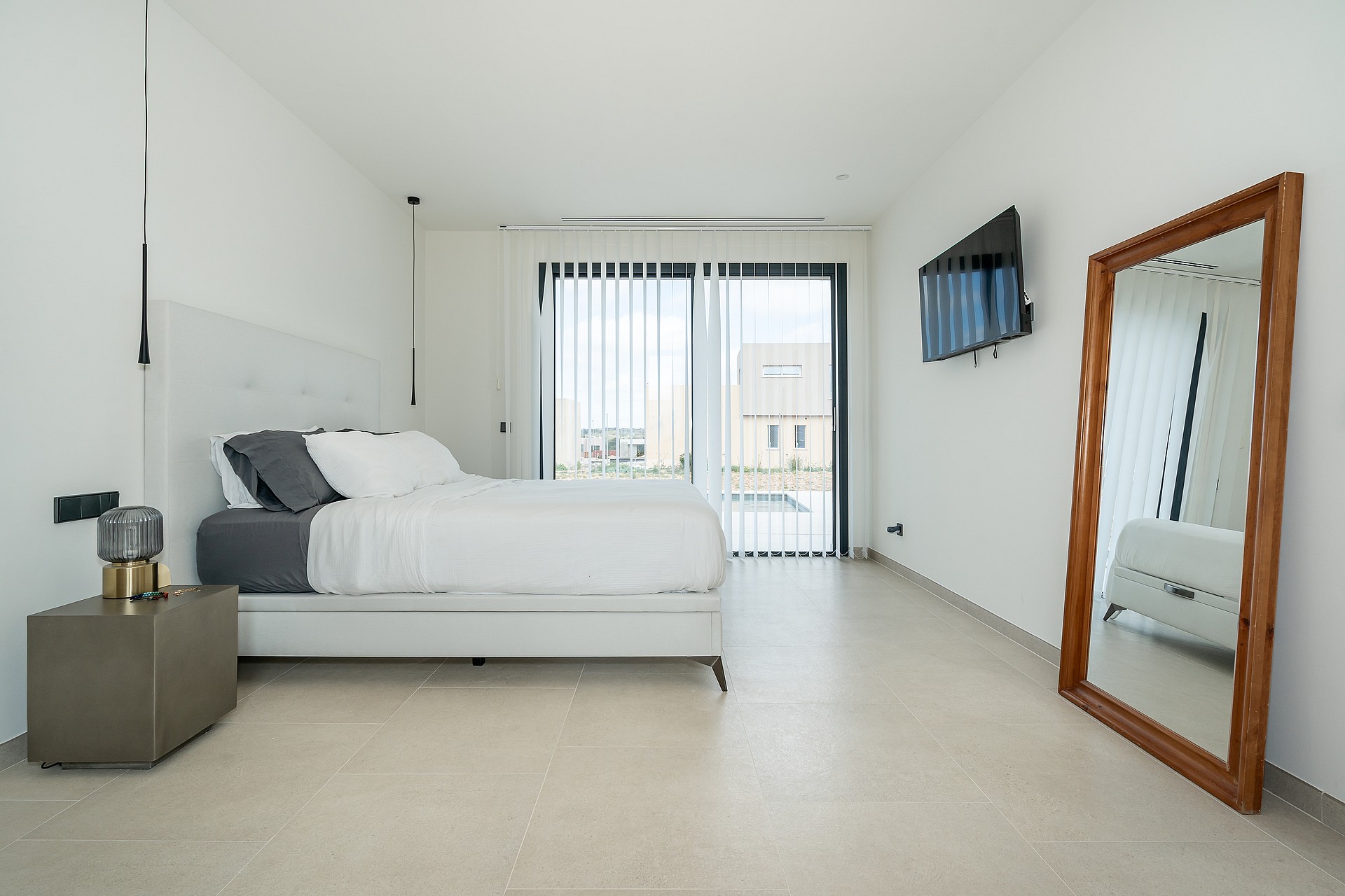 A minimalist bedroom featuring a white queen bed, light stone flooring, and a large wooden-framed floor mirror.