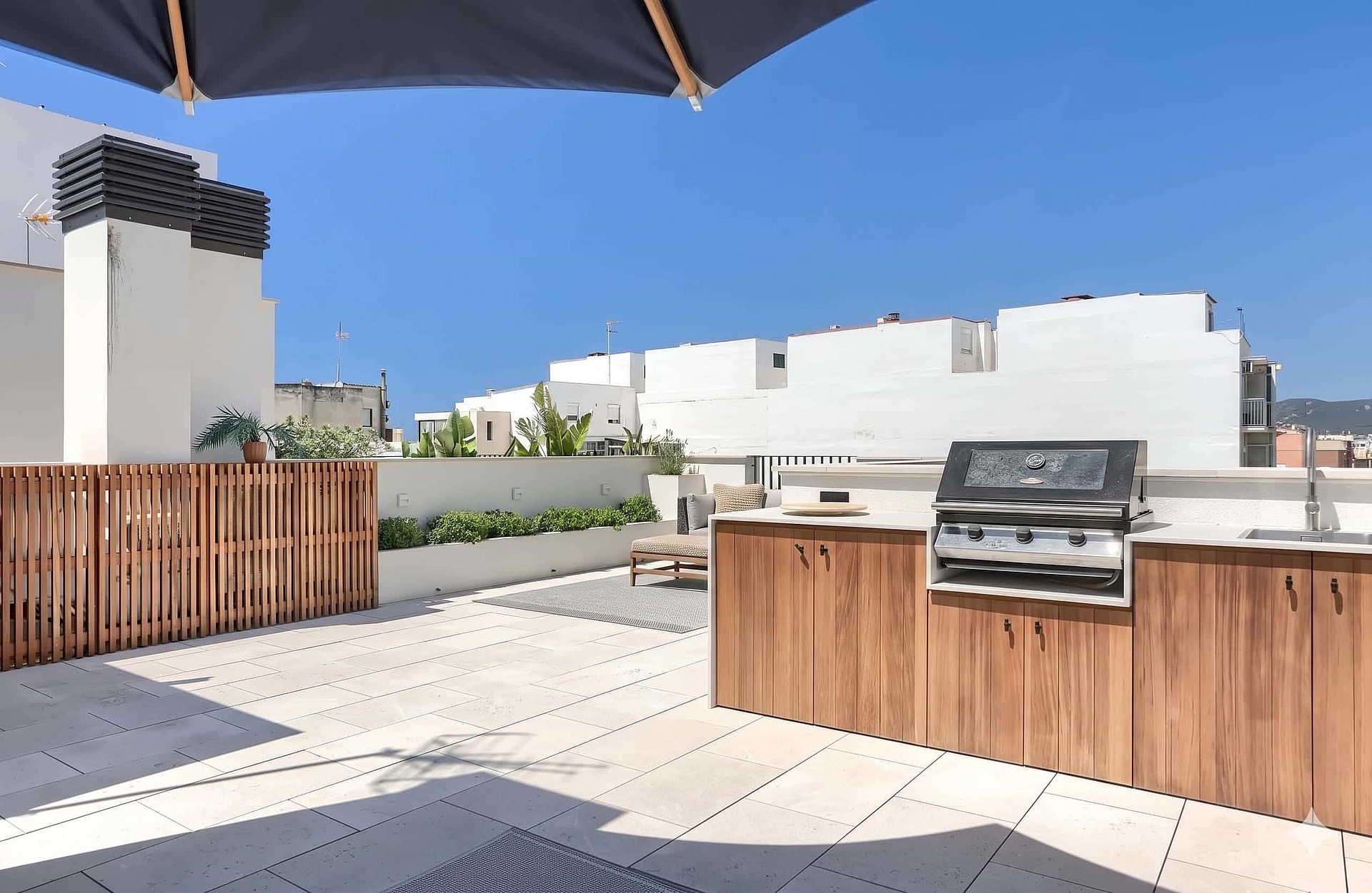 A rooftop terrace featuring a wooden outdoor kitchen, barbecue grill, and stone flooring under a clear blue sky.