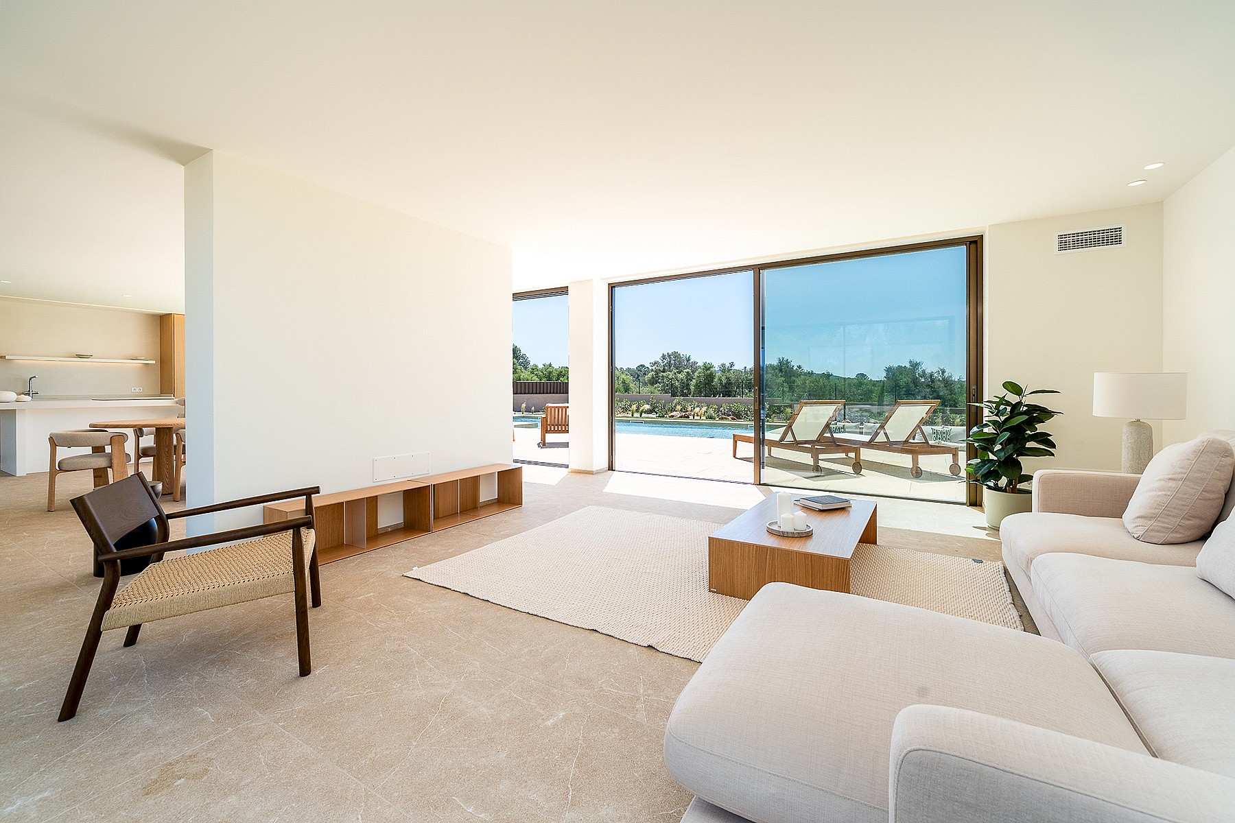 Bright open-plan living room featuring neutral sofas, a woven wood armchair, and floor-to-ceiling glass doors opening to a pool deck.