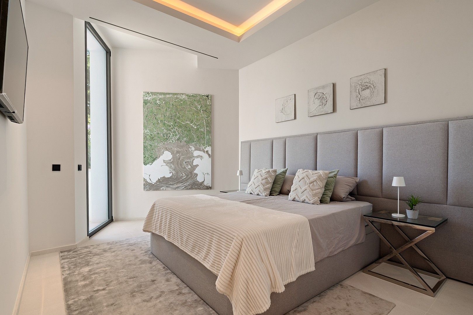A minimalist bedroom featuring a king-sized bed with a tall grey upholstered headboard, a textured area rug, and cove lighting.