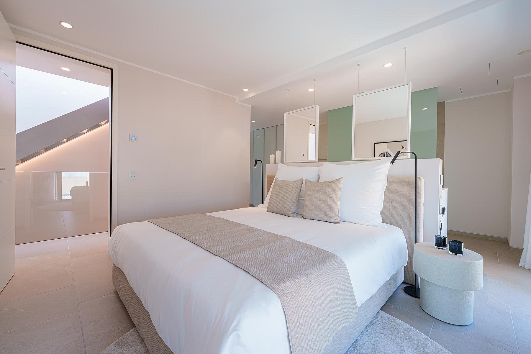 Dormitorio moderno con cama central, ropa de cama blanca y tonos beige sobre paredes de color claro.