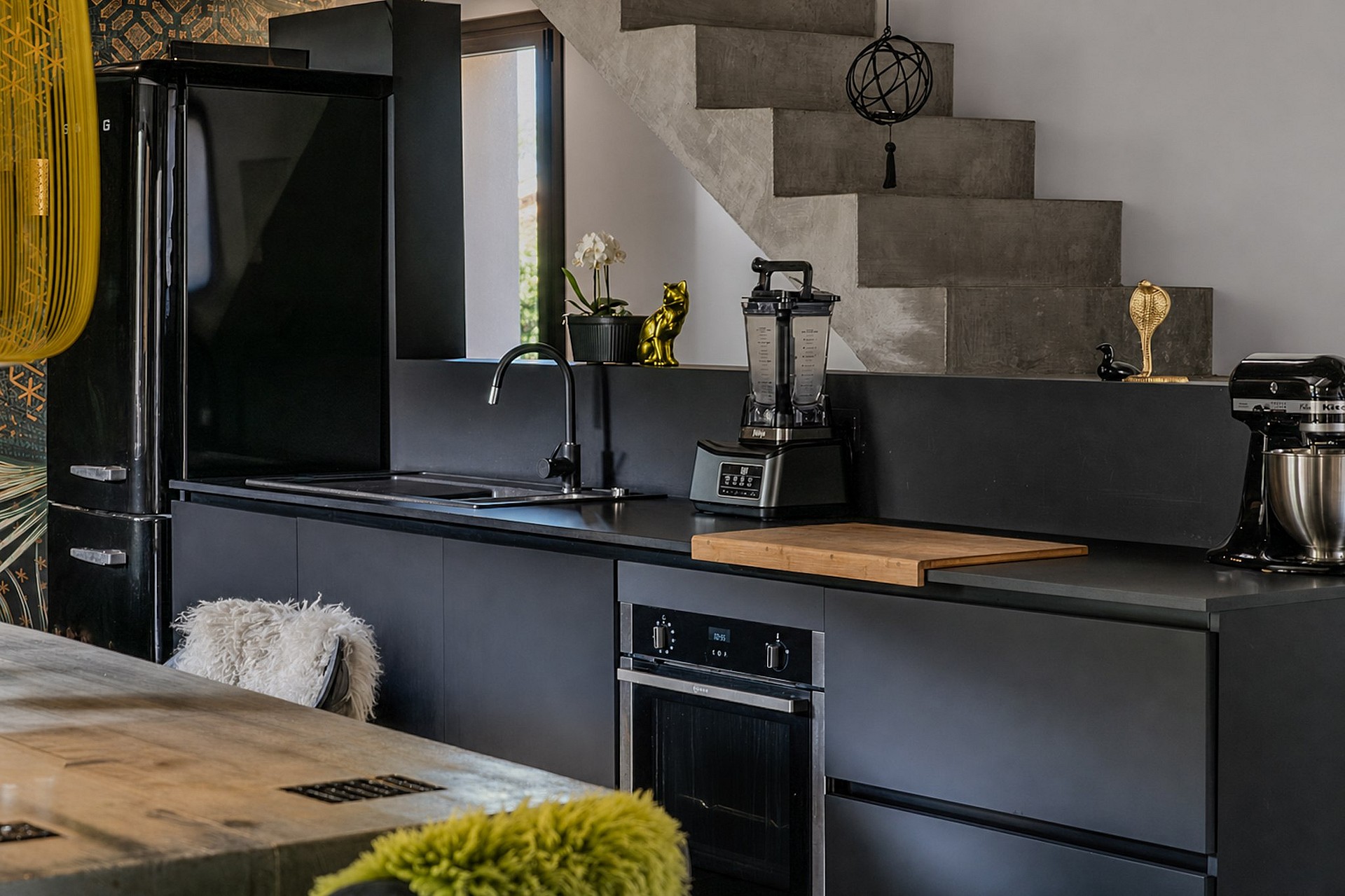Modern black kitchen with matte cabinetry, wooden island countertop, and stone staircase in the background.