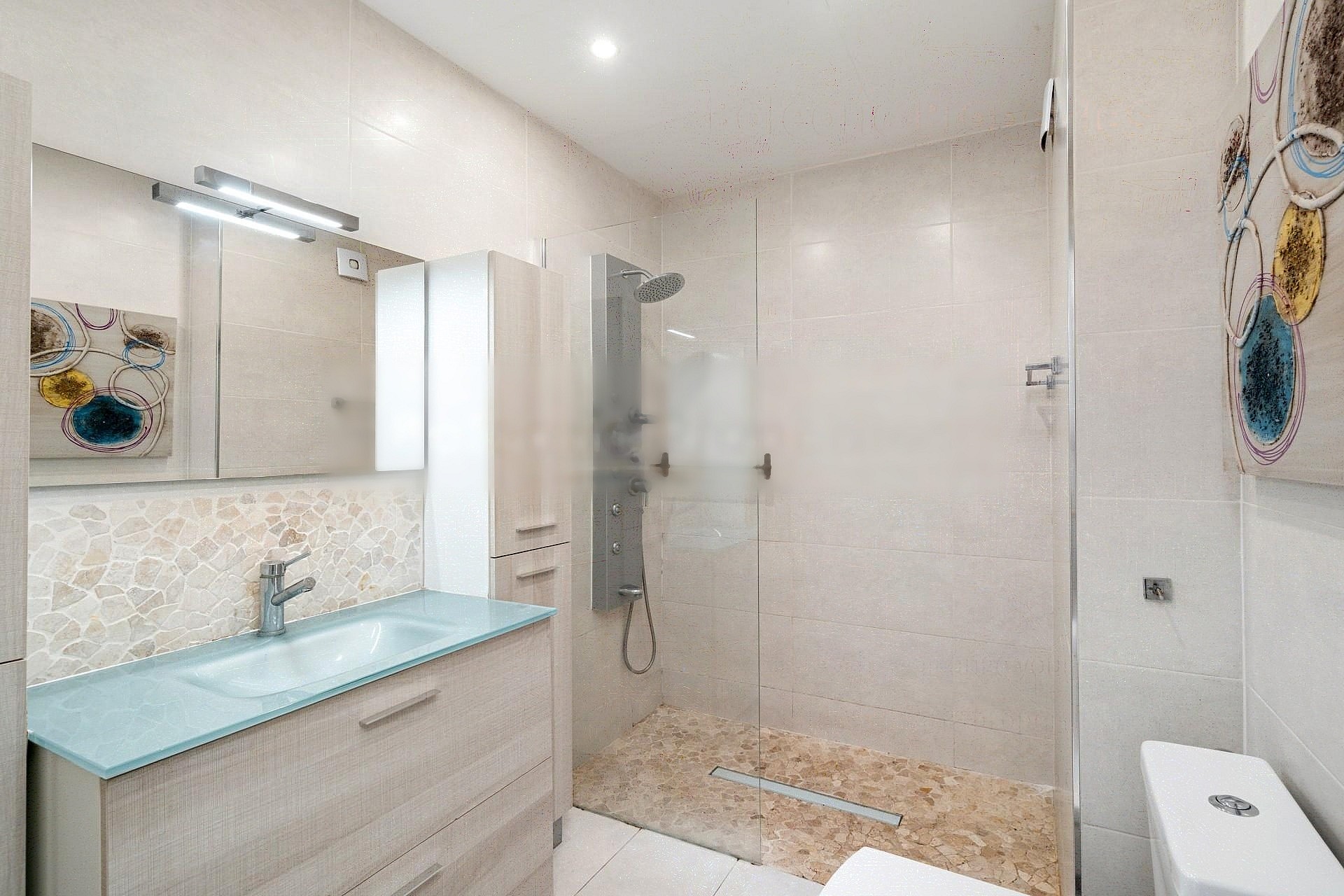 A contemporary bathroom featuring a light-toned wooden vanity, glass sink, and a walk-in shower with textured stone floor tiles.