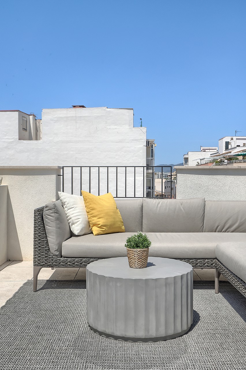 A rooftop terrace in El Molinar featuring a grey modular sofa with yellow cushions, a round fluted concrete table, and a patterned rug against a bright sky.