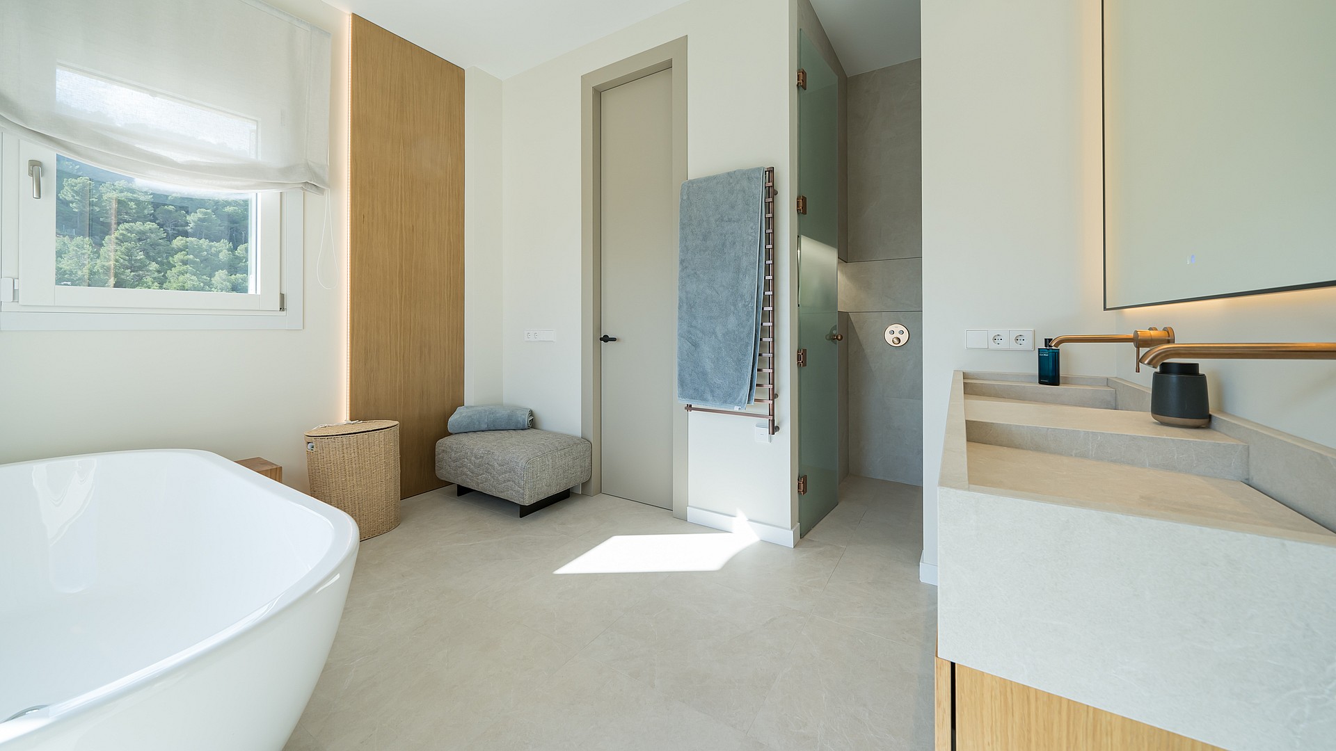 A bright bathroom featuring a white freestanding bathtub, a stone double vanity with bronze fixtures, and warm wood accents.
