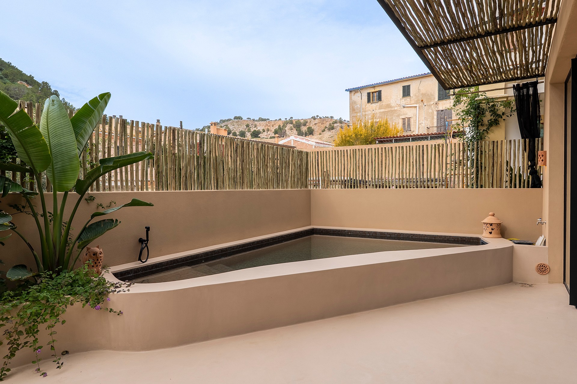 A small corner swimming pool on a terrace with beige stucco walls, bamboo fencing, and a reed shade cover.