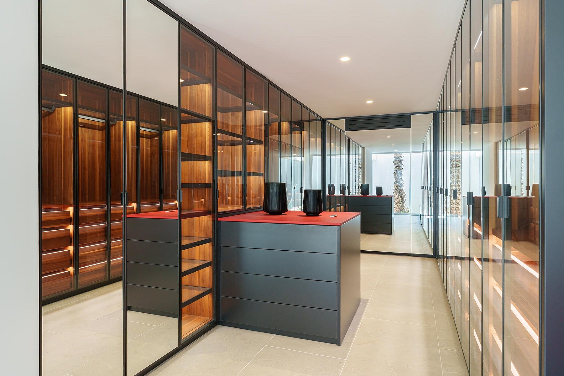 A walk-in dressing room featuring dark wood shelving enclosed by glass panels, with a central dark matte island unit on a light stone floor.