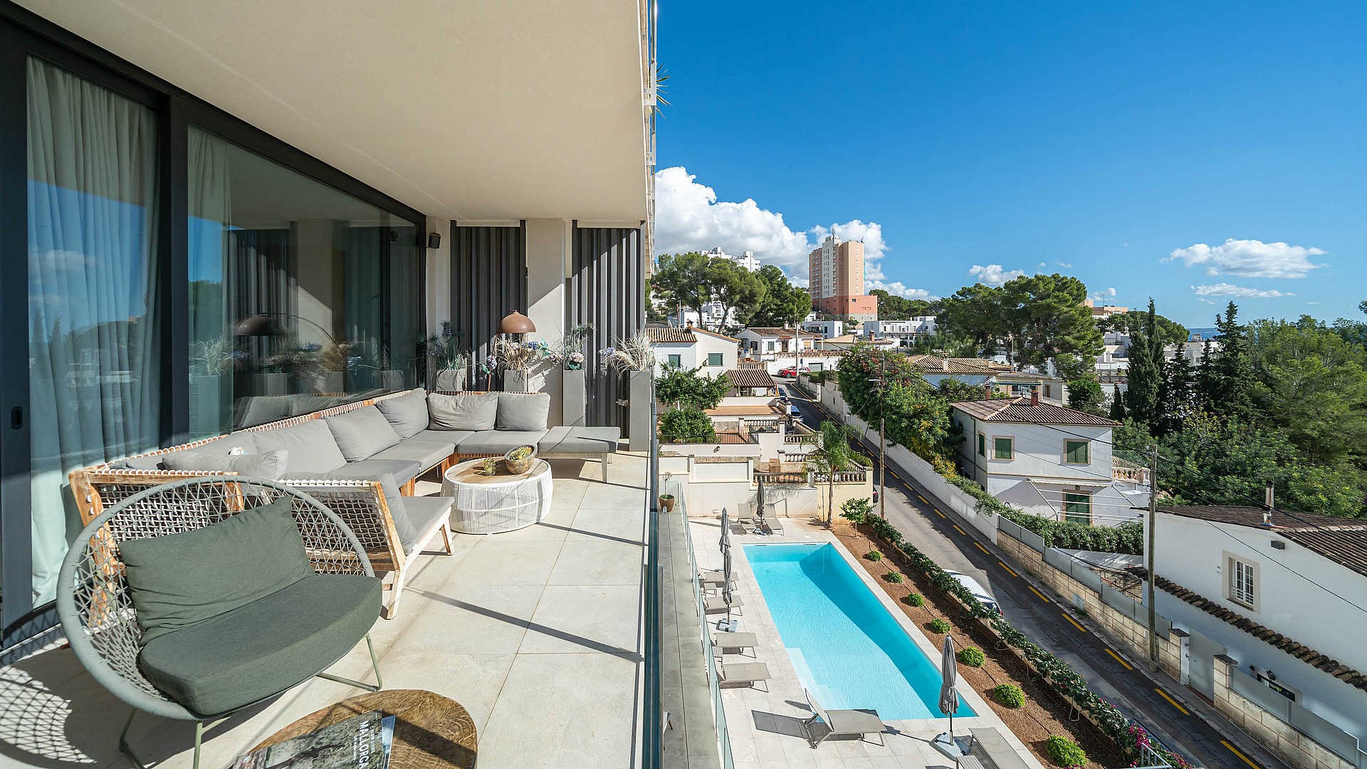 A covered terrace with grey sectional seating overlooks a rectangular swimming pool surrounded by a paved garden in a suburban setting.