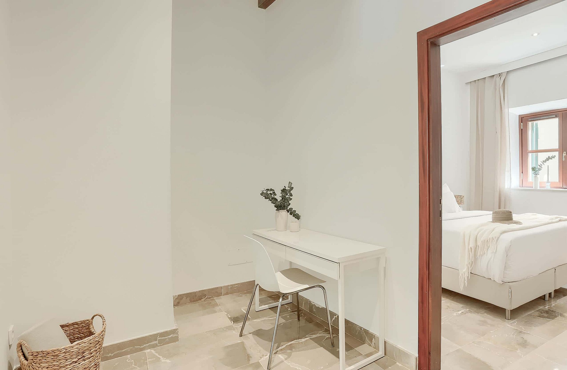 A small minimalist study area with a white console desk and chair, located in an alcove next to a doorway showing a bedroom.