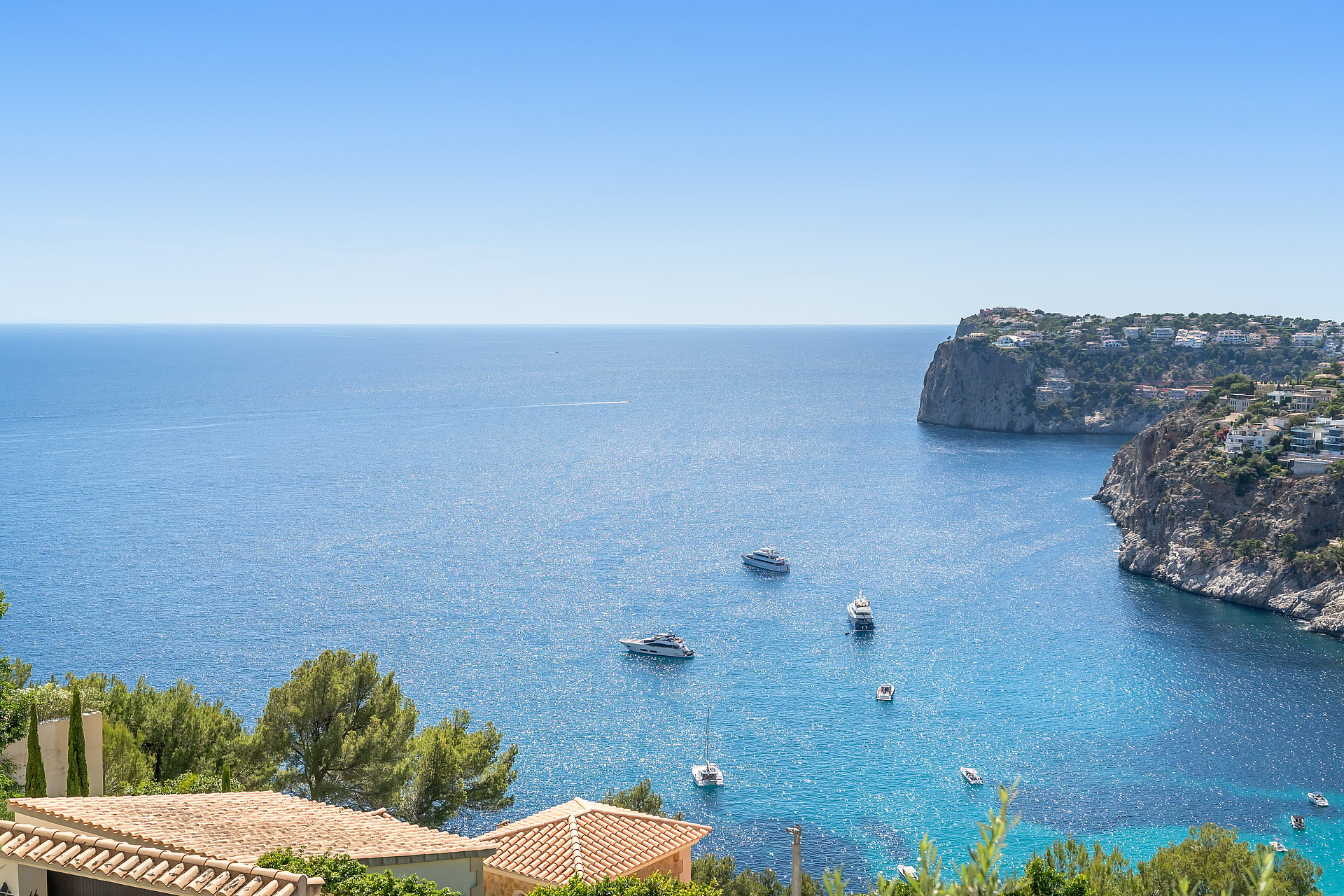 Elevated coastal view showing anchored yachts in a bright blue bay surrounded by rugged rocky cliffs and green hills.