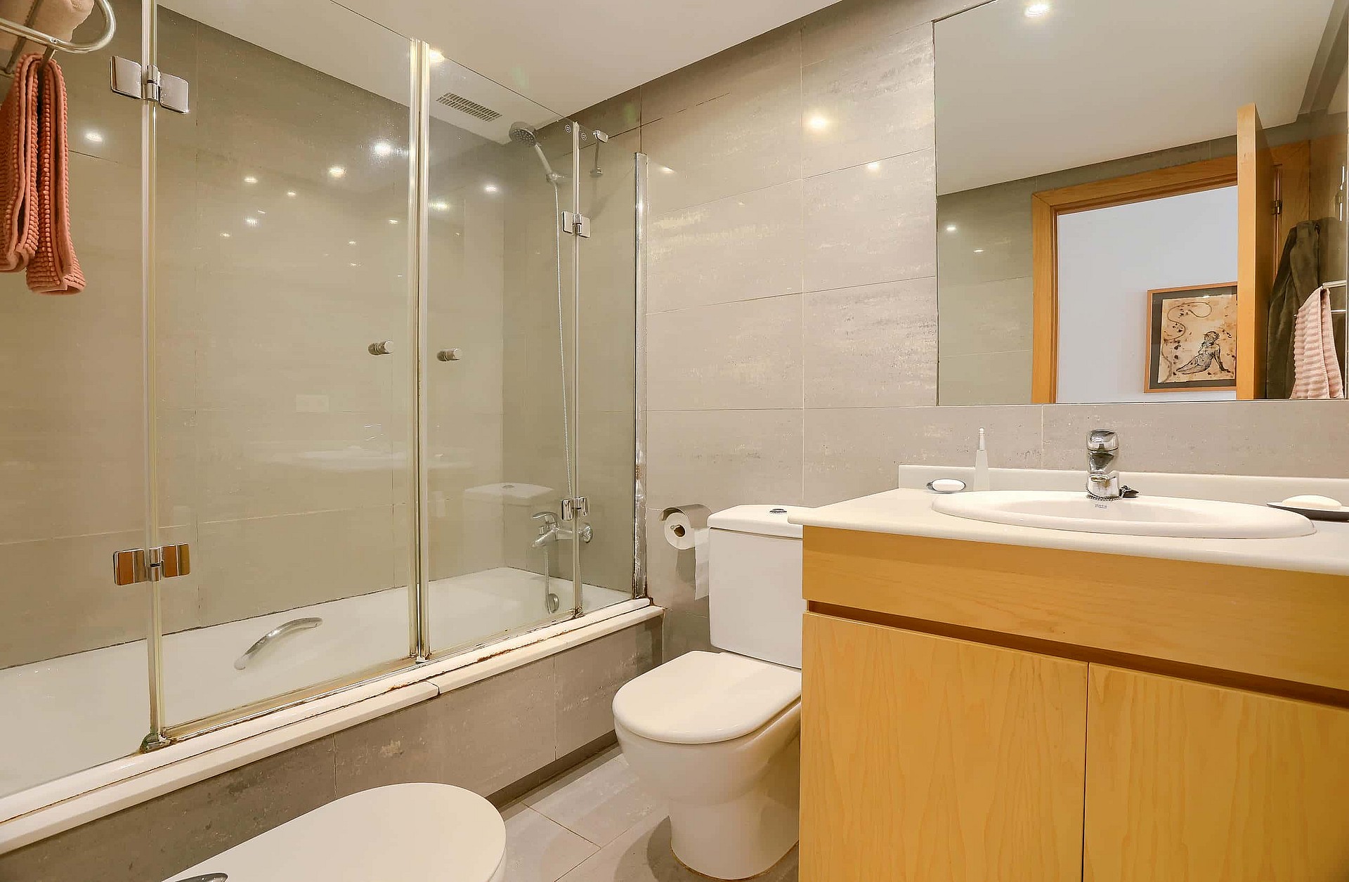 A bathroom featuring a light wood vanity, a wall-mounted mirror, a glass-enclosed bathtub, and neutral wall tiles.