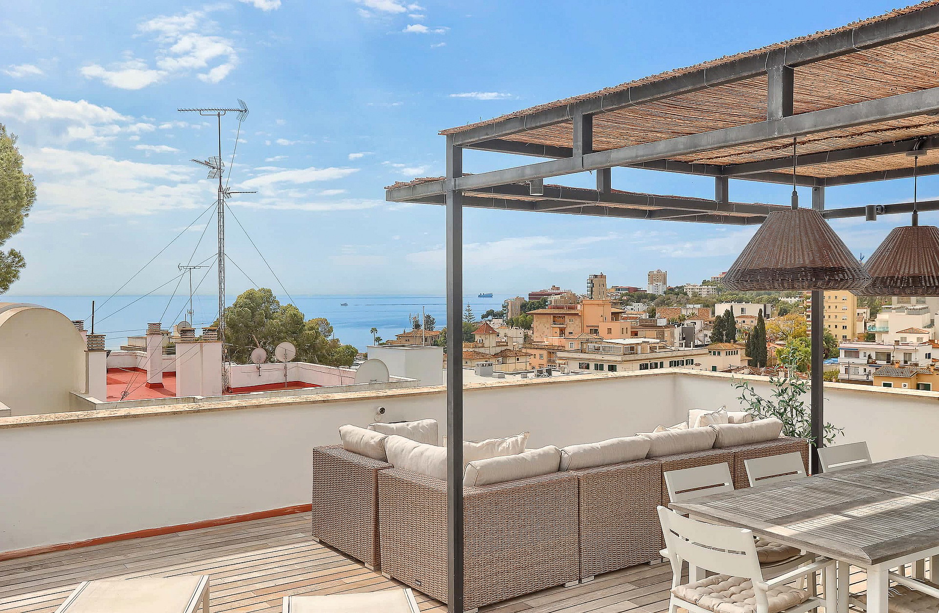 A rooftop terrace featuring a wicker sofa, dining set, and a pergola against a backdrop of the Mediterranean Sea and San Agustin.
