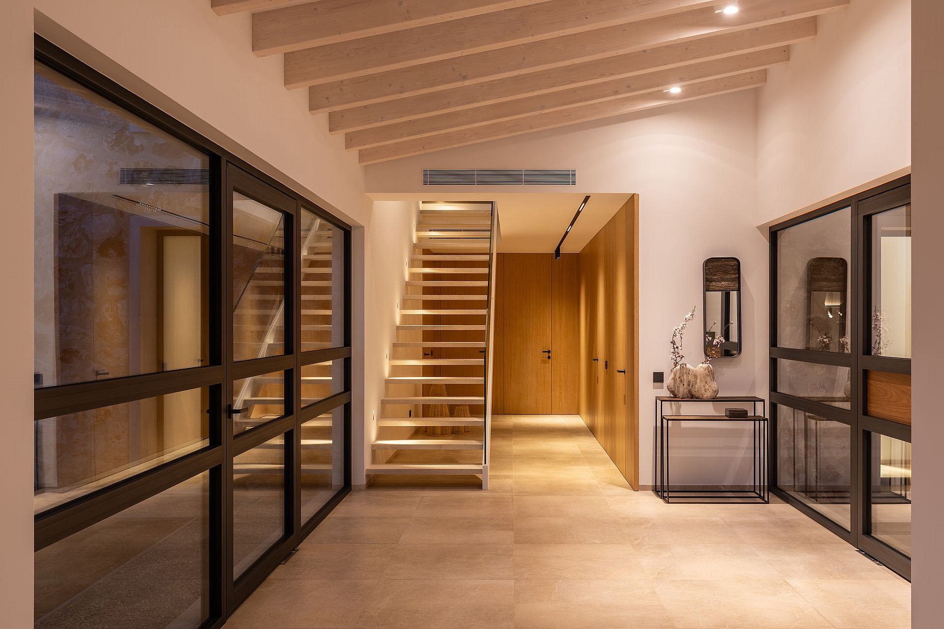 Interior entrance hall of a modern finca featuring a floating wooden staircase, light-toned stone flooring, and timber ceiling beams.