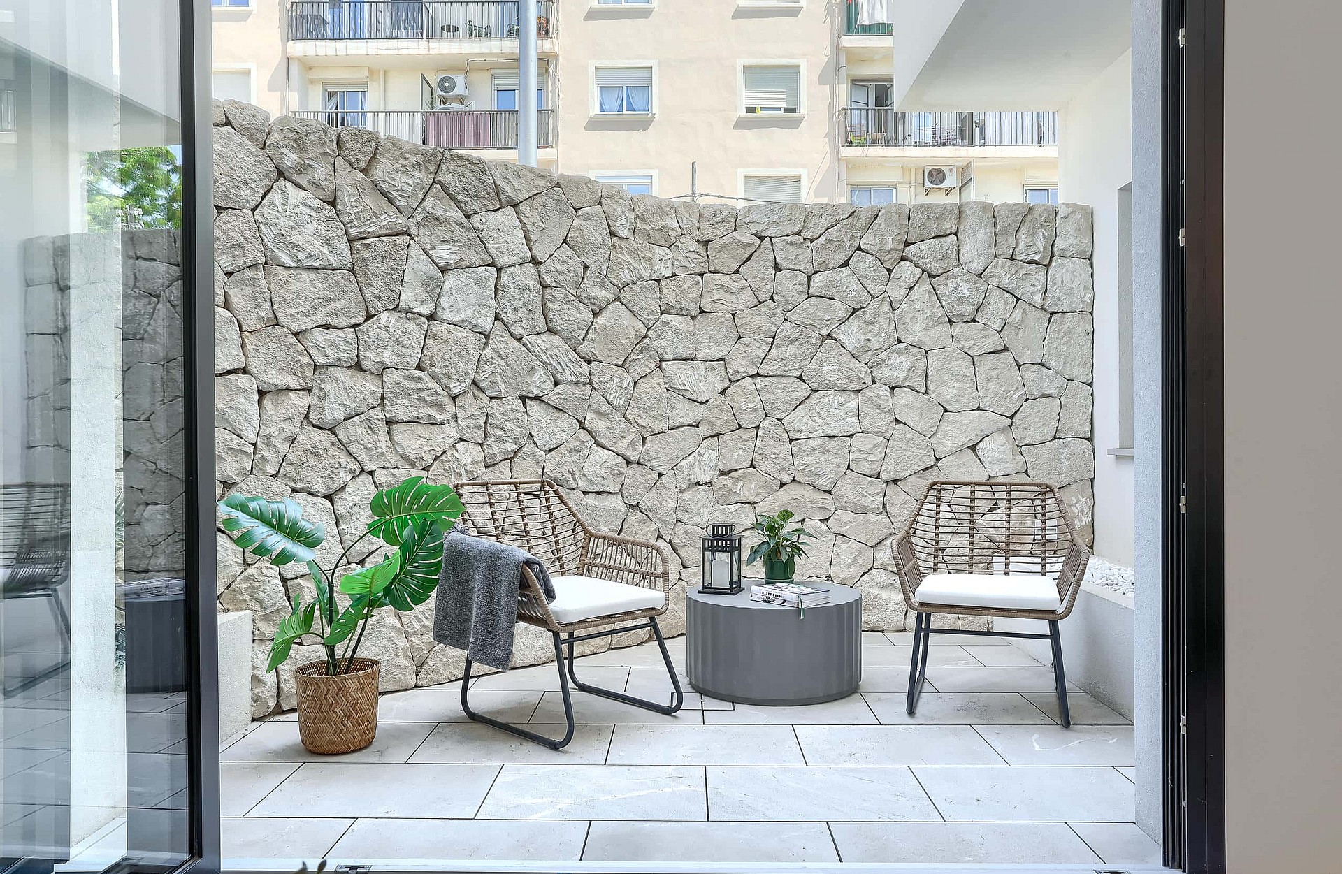 A private outdoor patio featuring a high dry-stone boundary wall, light-toned stone paving, and two rattan chairs with a small table.