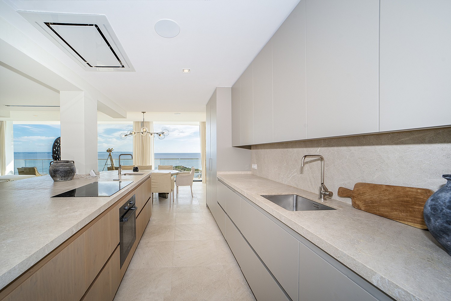 Galley kitchen with light grey handleless cabinets, stone countertops, and built-in appliances, opening to a dining area overlooking the sea.