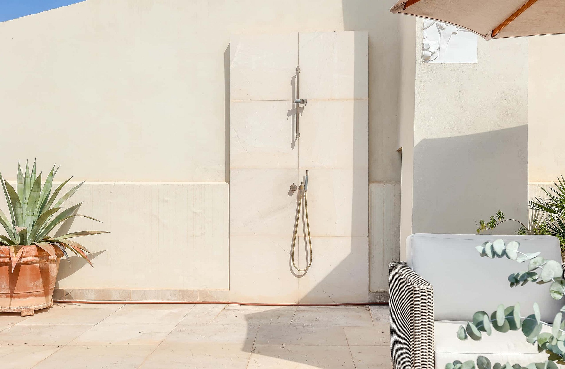 An outdoor shower fixture mounted on a light stone wall on a sunny terrace with a large potted agave plant.