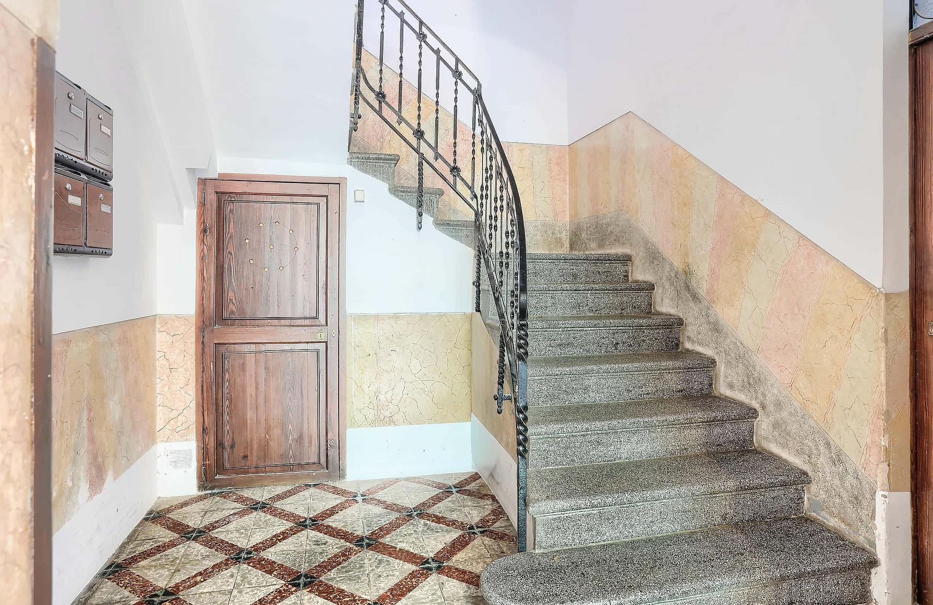 A staircase with terrazzo steps and a black wrought-iron railing in a residential building with a decorative patterned floor.