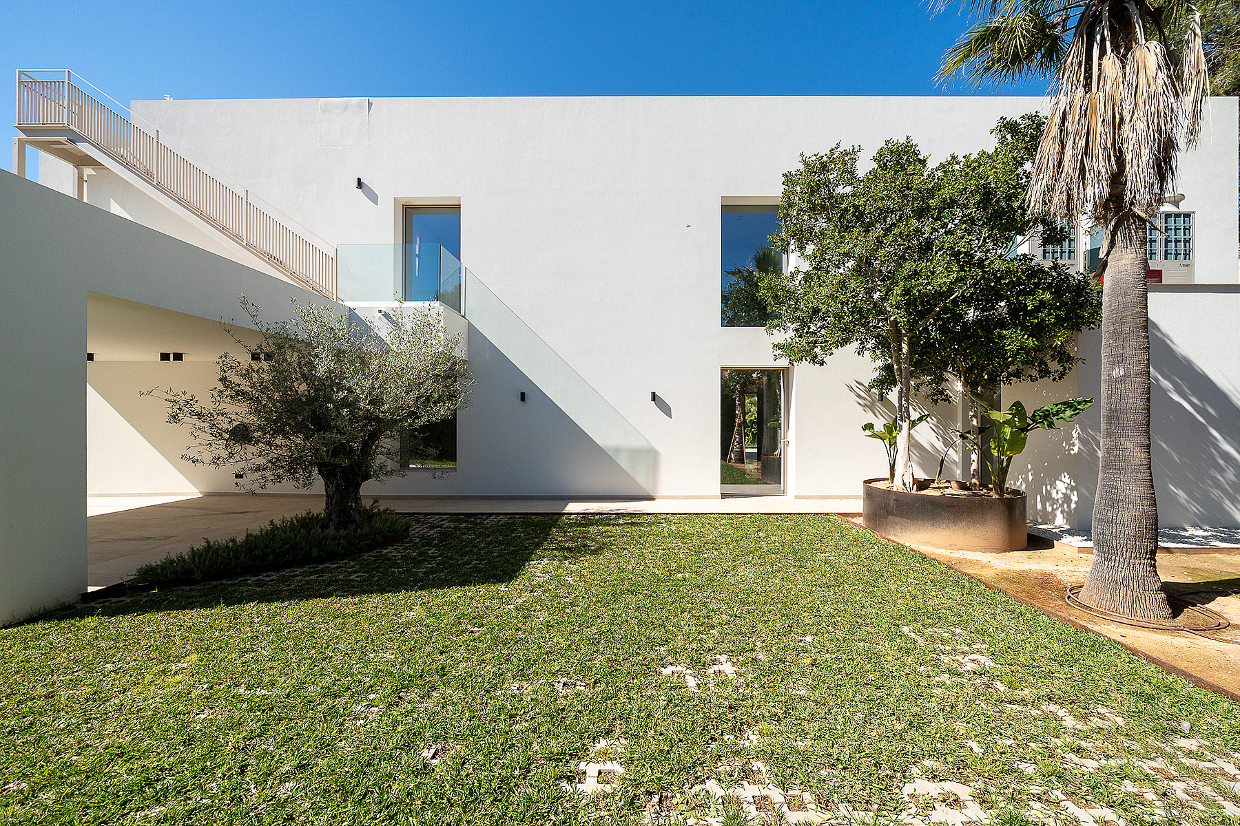 Modern white minimalist villa exterior featuring a flat roof, large rectangular windows, and a manicured green lawn with mature trees.