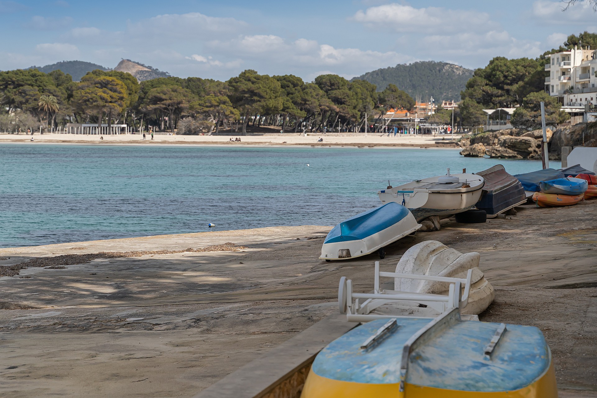 Small boats parked on a stone ramp facing a calm turquoise bay with a sandy beach and dense pine forest in the distance.