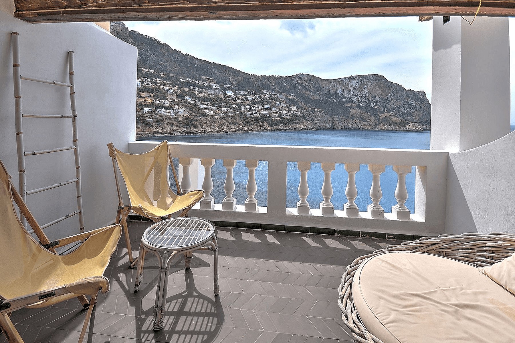 Covered terrace with stone floor, two yellow folding chairs, and a rattan lounge seat overlooking the ocean and mountains.