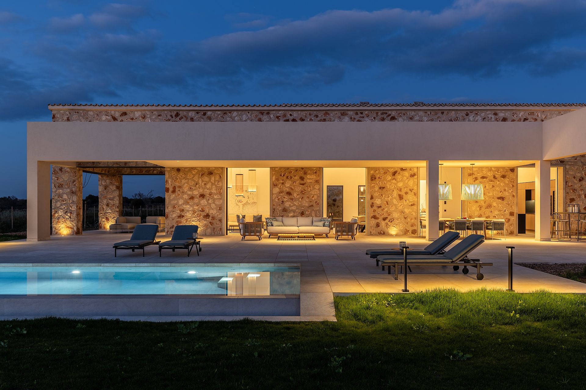 Exterior view of a stone finca with a pool and terrace illuminated at twilight, featuring outdoor furniture and lawn.