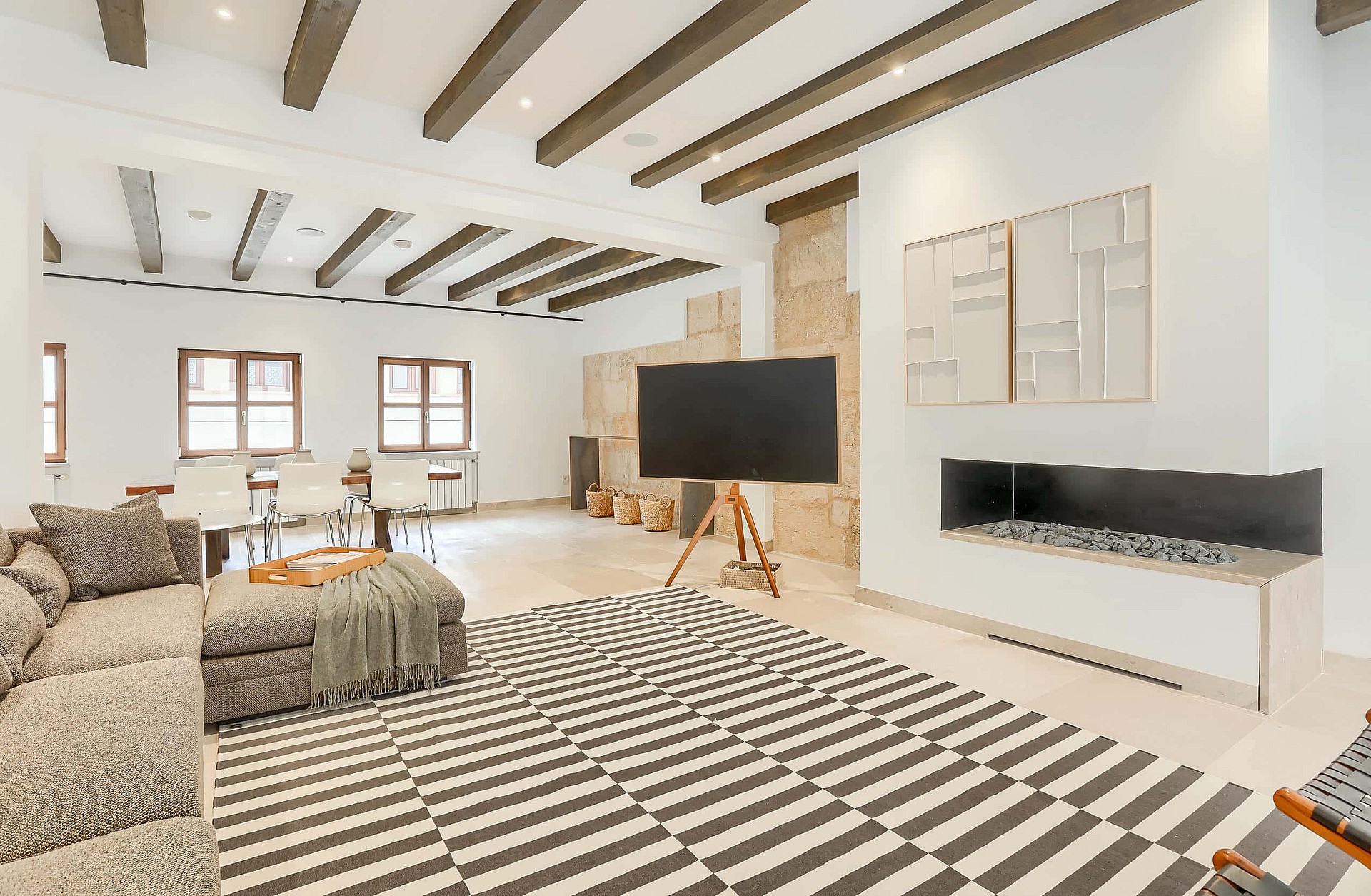 Wide angle view of an open-plan living room featuring exposed dark wood ceiling beams, white walls, a stone-surround fireplace, and a large patterned area rug.