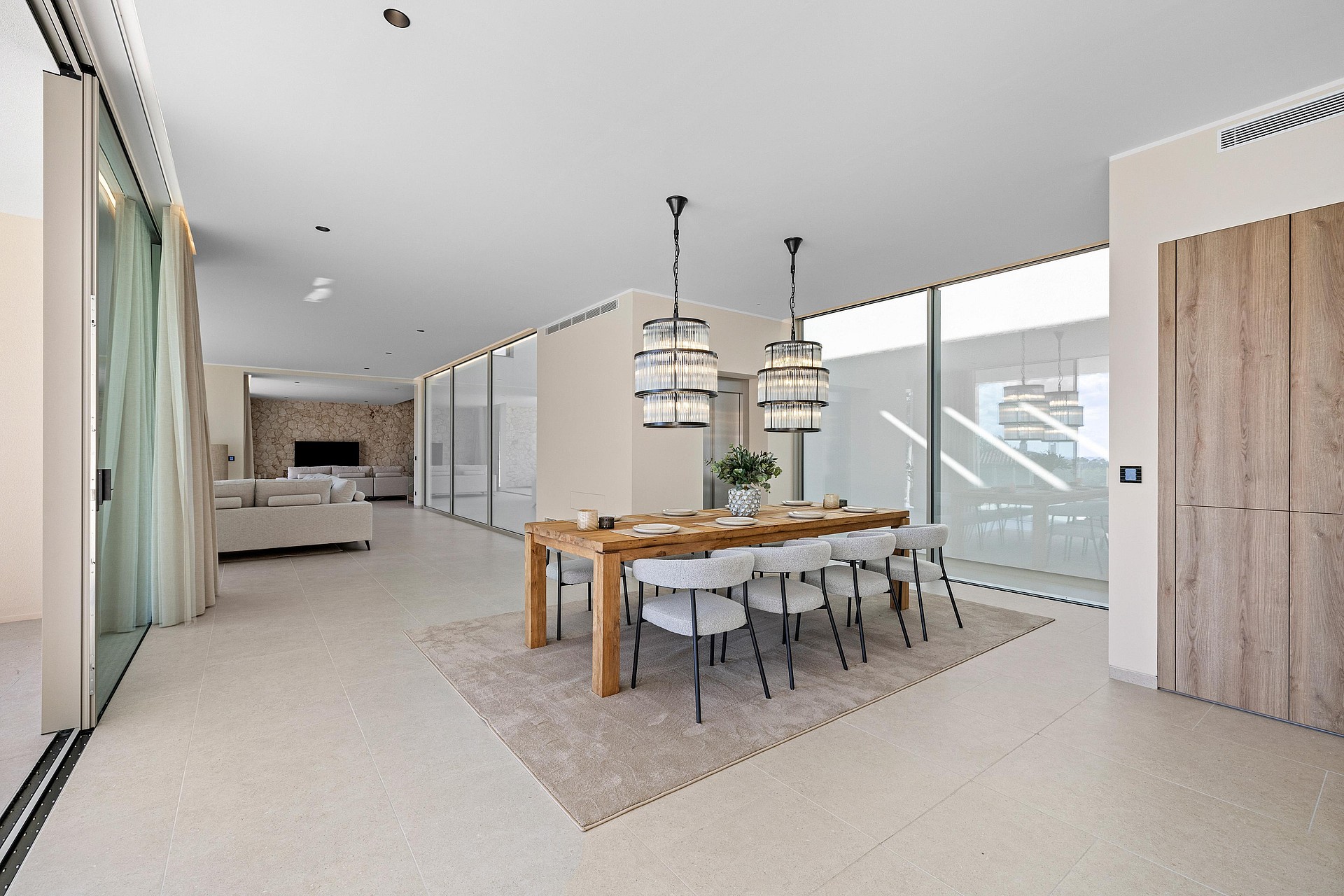 Open-plan living and dining area featuring floor-to-ceiling windows, a wooden dining table, fabric chairs, and designer lighting.