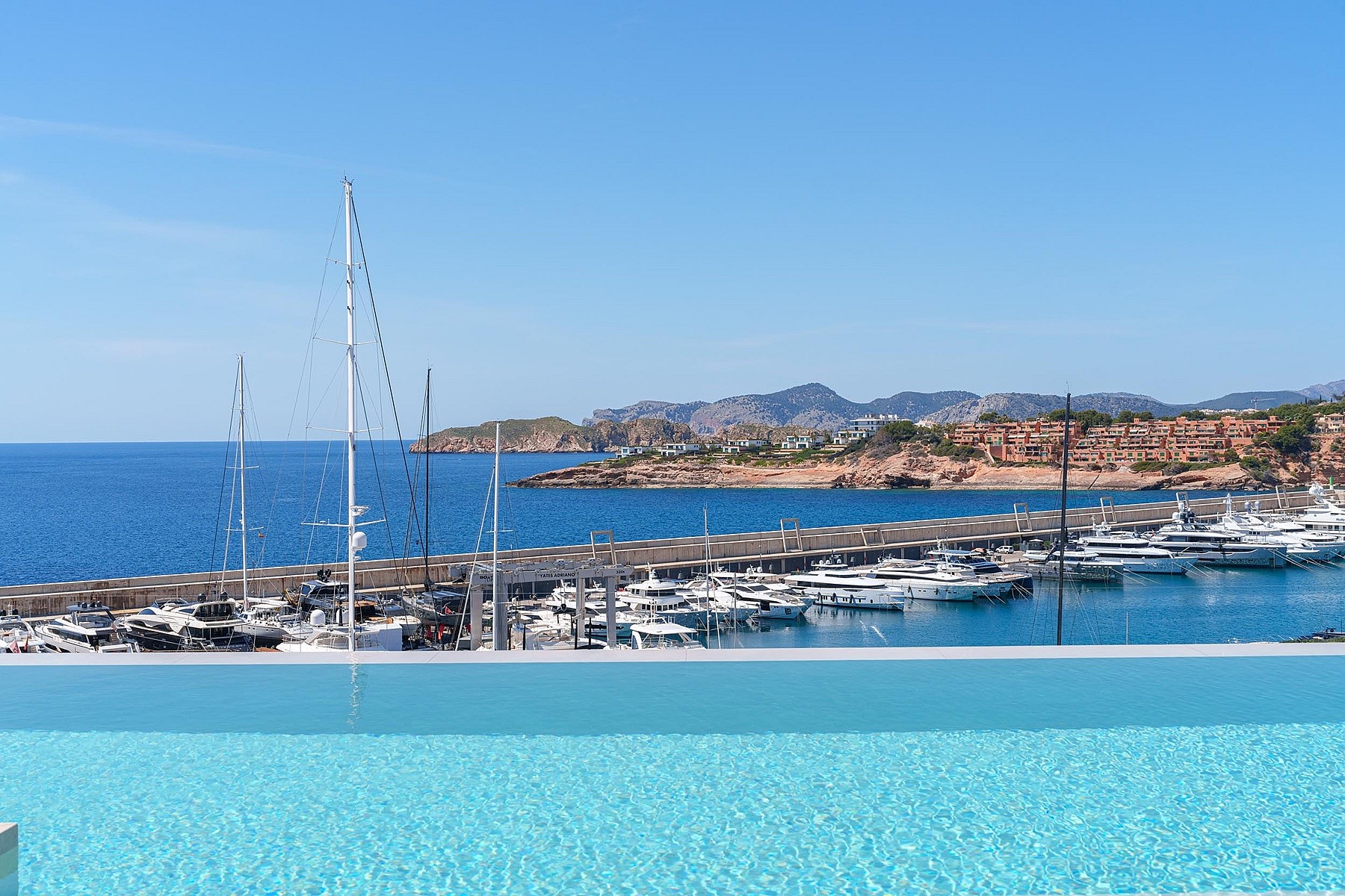 An infinity pool edge looking out towards the Port Adriano marina with yachts and the Mediterranean sea under a clear sky.