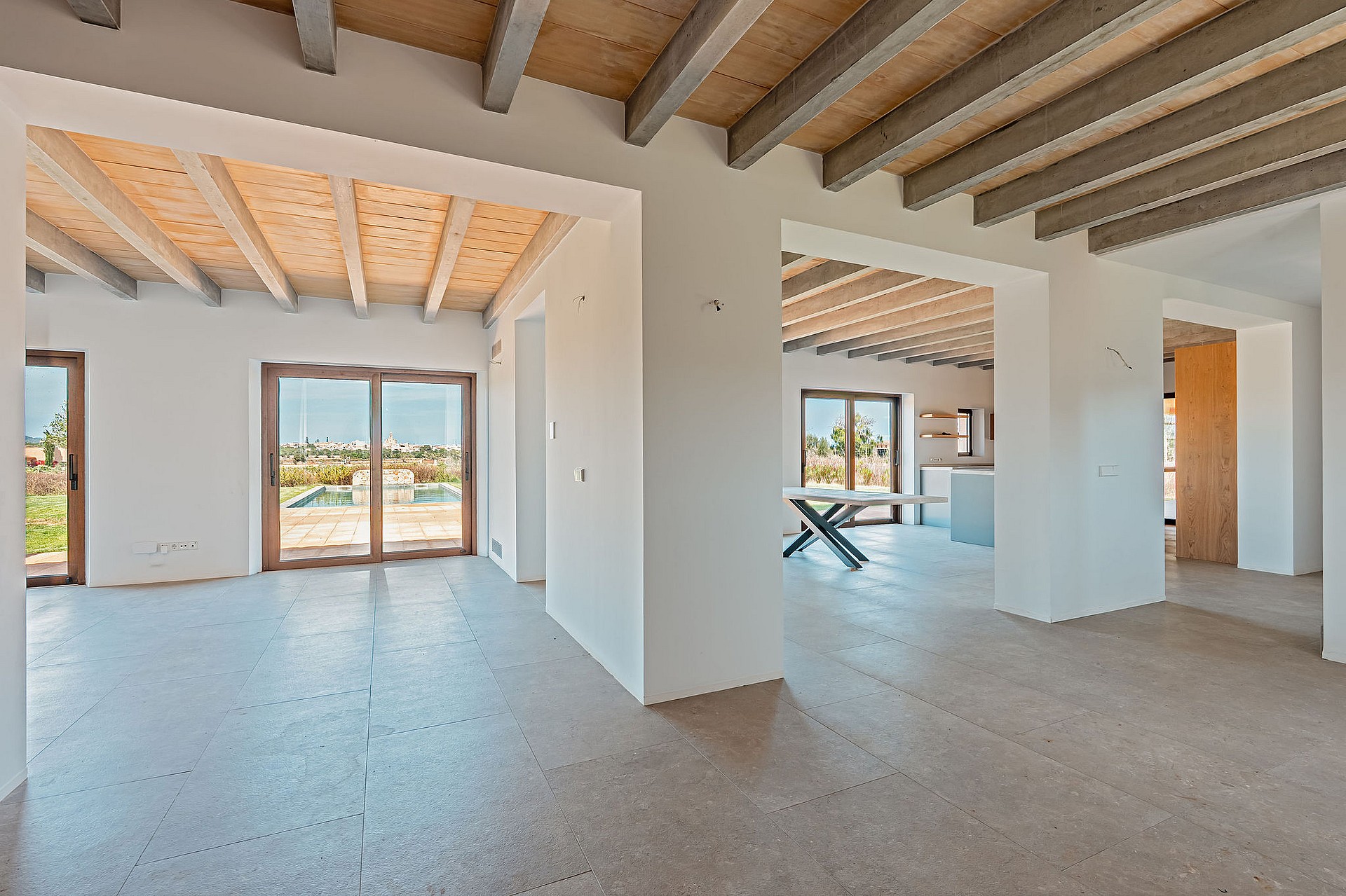 Empty open-plan living room in a finca with white walls, exposed wood ceiling beams, and stone tile floors.