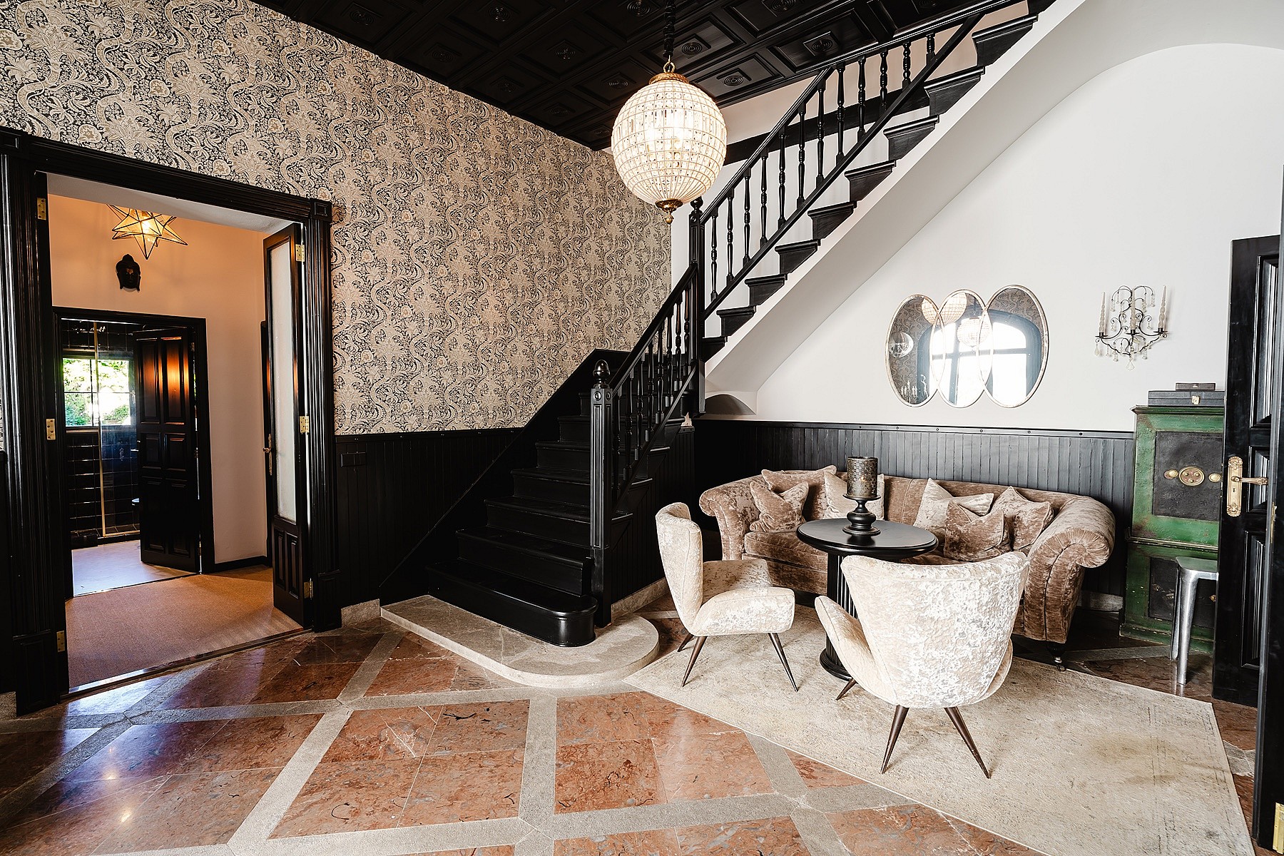 Entrance foyer featuring a dark wood staircase, patterned wallpaper, a crystal pendant light, and a seating area with ornate chairs.