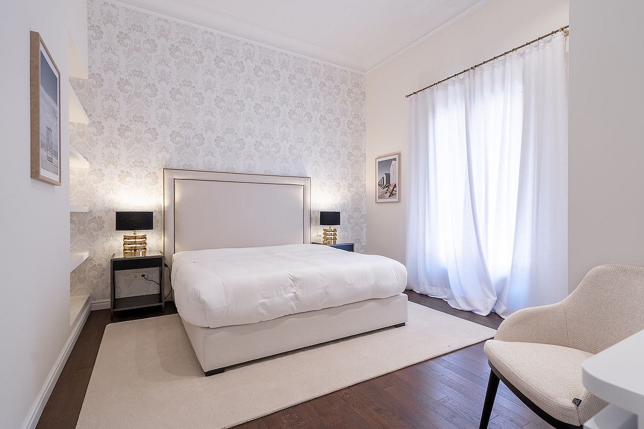 A bright bedroom featuring a queen bed, beige rug, white patterned wallpaper, and sheer curtains over a window.