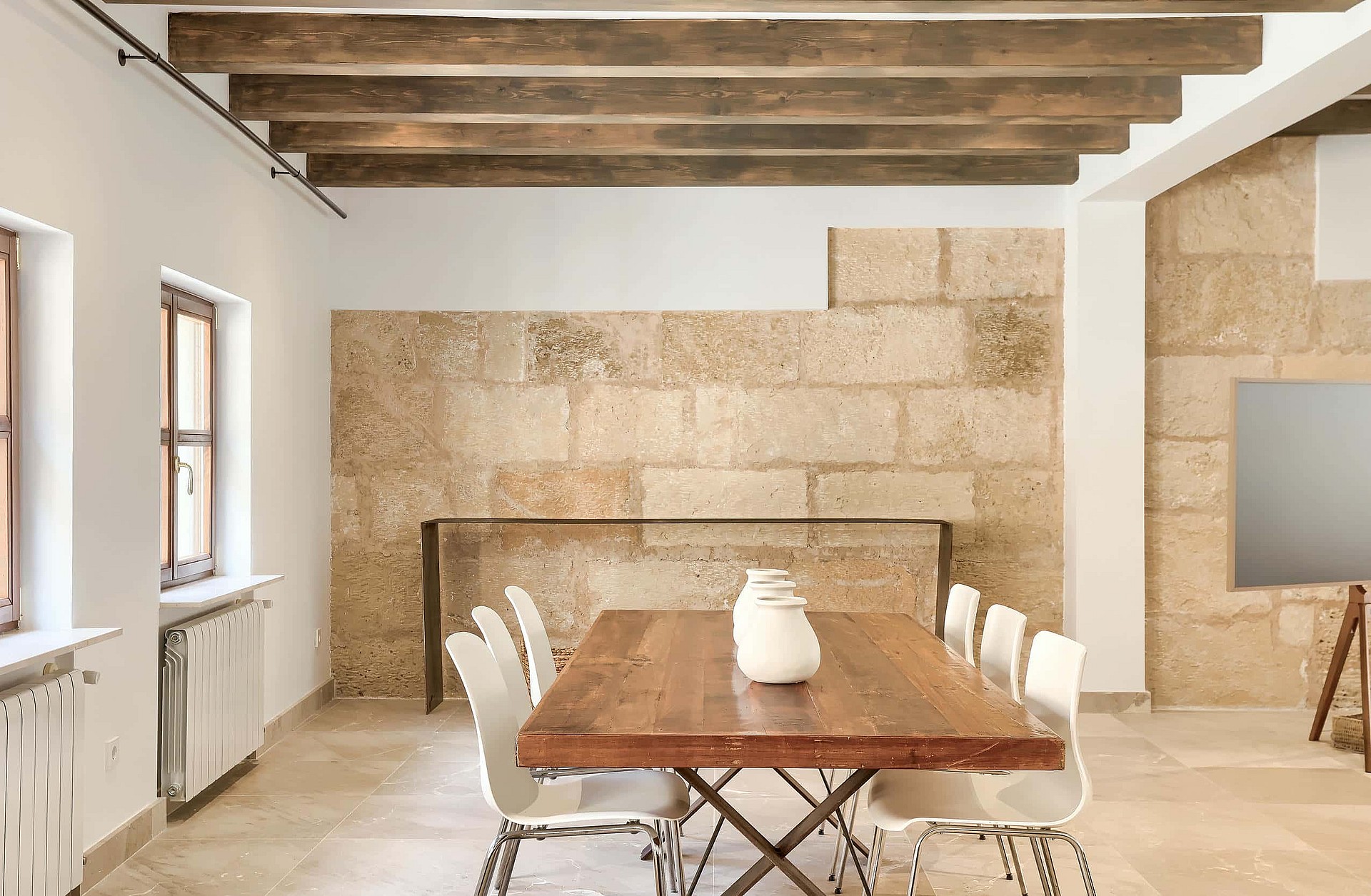 Dining area with a long wooden table, modern white chairs, exposed timber ceiling beams, and textured stone block wall.